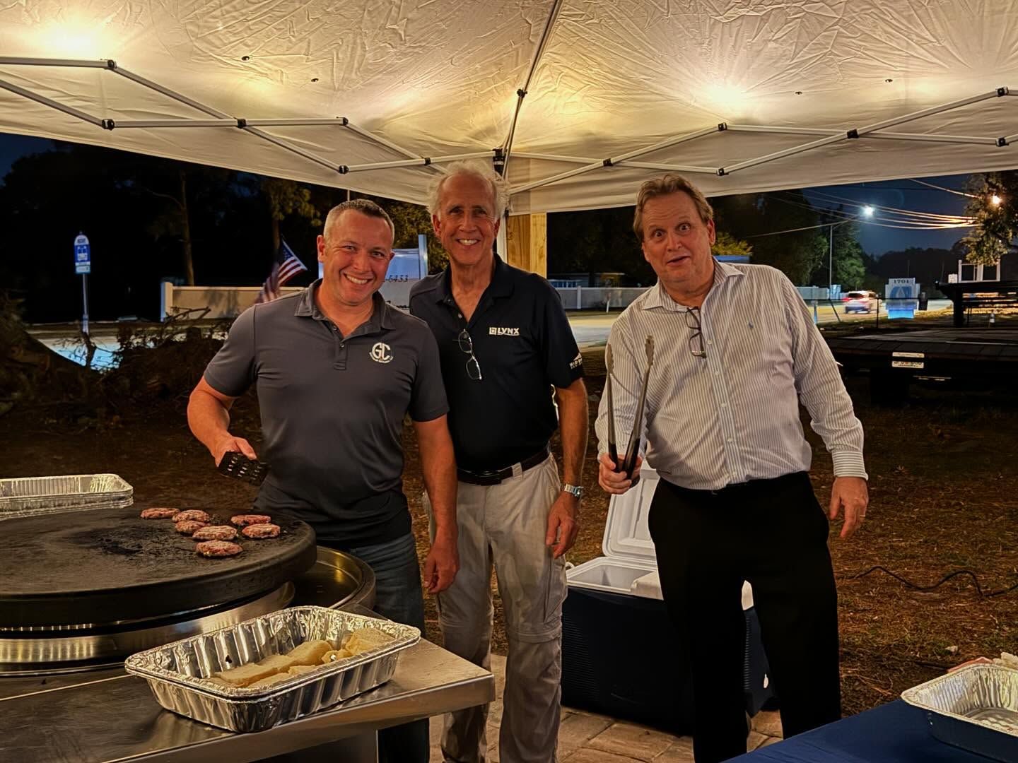 Three men grilling under a tent at night; one flips burgers.