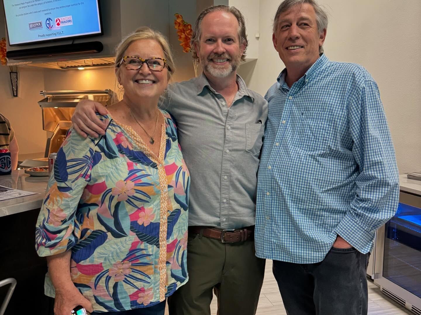 Three people smiling, posing together indoors. A woman with glasses and a colorful top, two men.