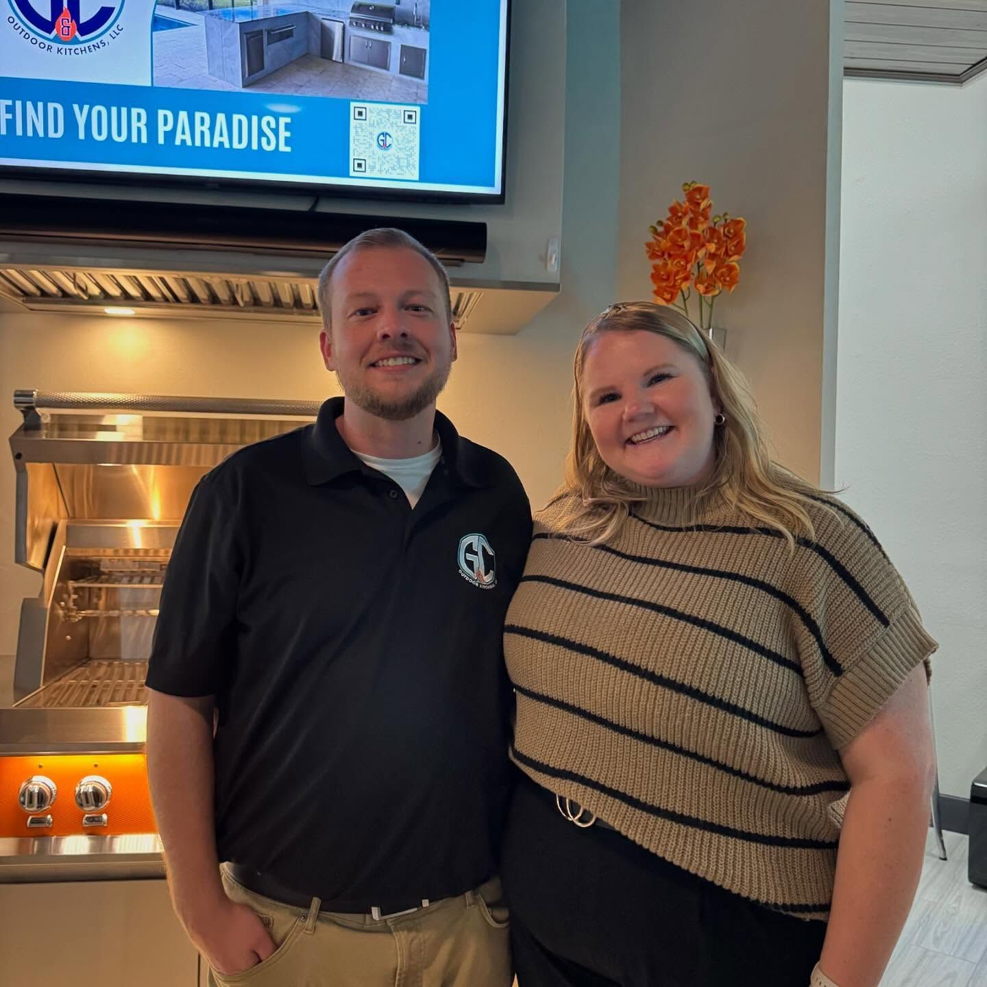 Two people smiling in front of an outdoor kitchen display. One wears a black shirt, the other a striped sweater.