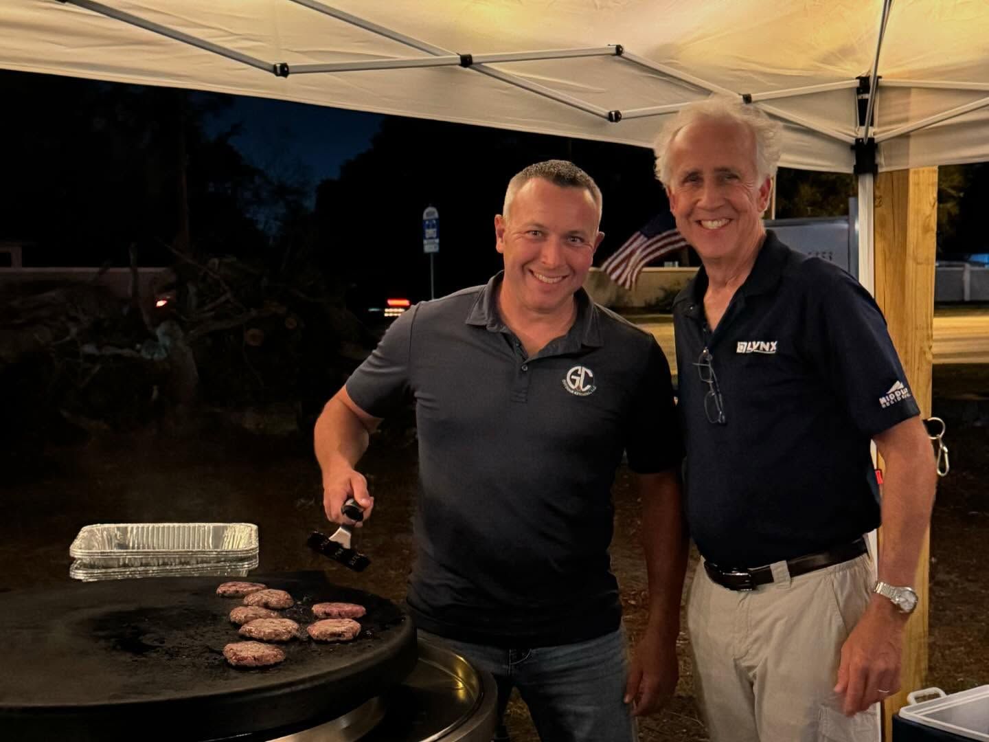 Two men grilling burgers at an outdoor event under a tent at night.