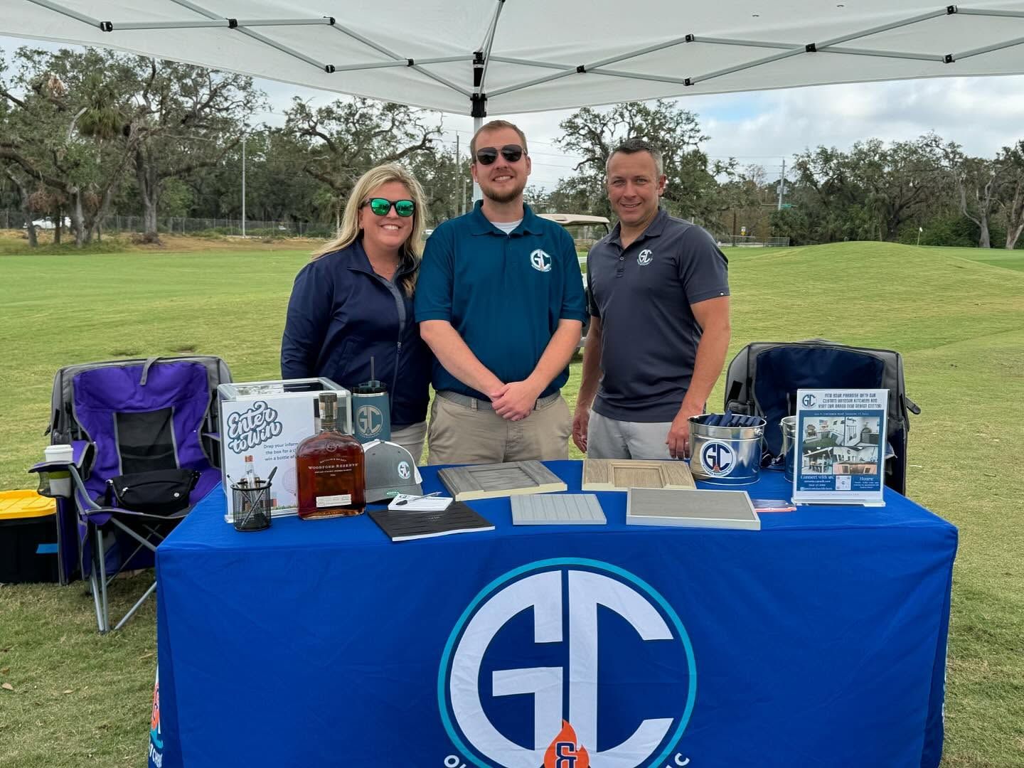 Three people stand behind a blue table at an outdoor event. They are under a white canopy, a grassy field is in the background. The table has the logo 