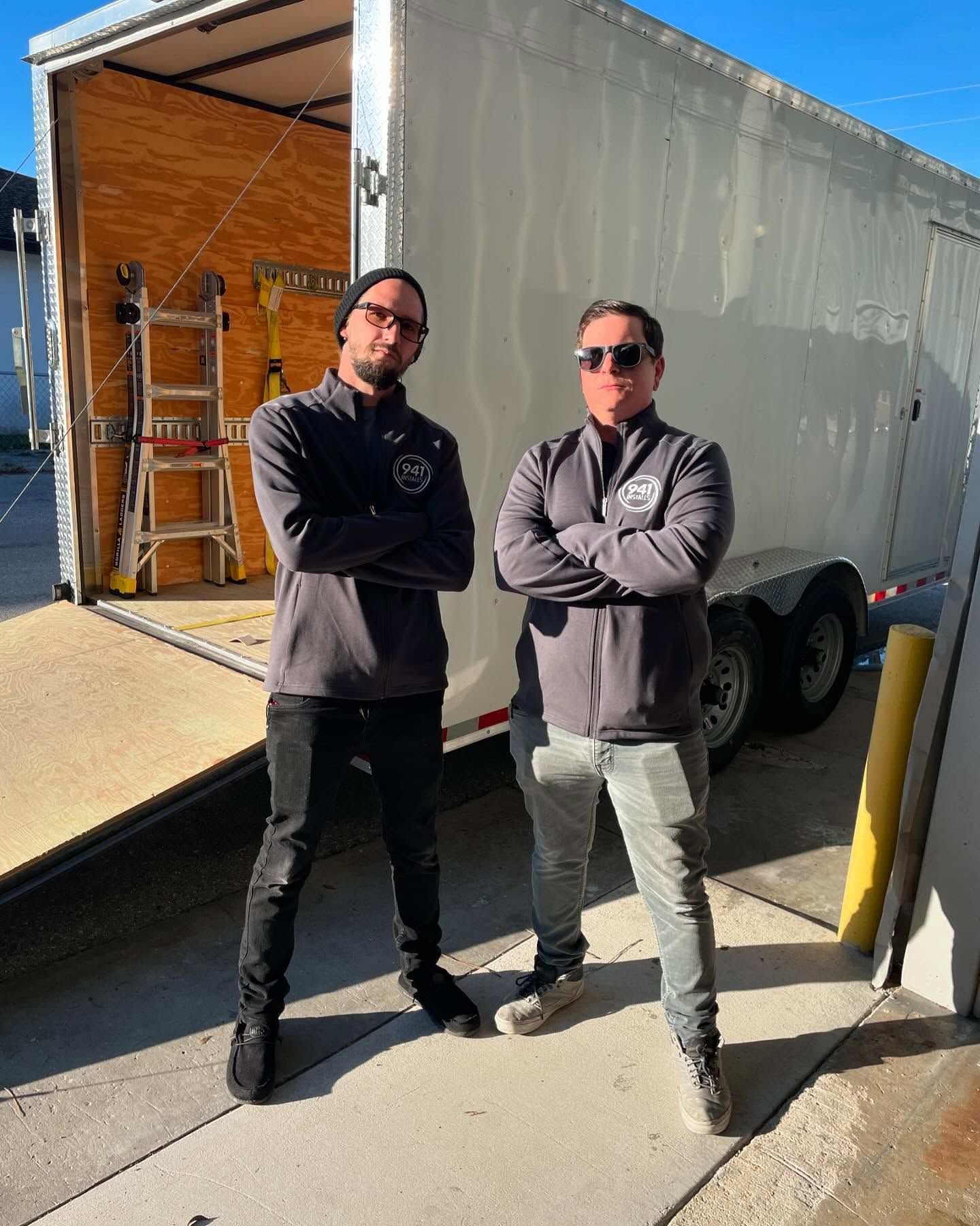 Two people stand with arms crossed near a trailer. One wears black pants, the other gray. Sunny outdoor setting.