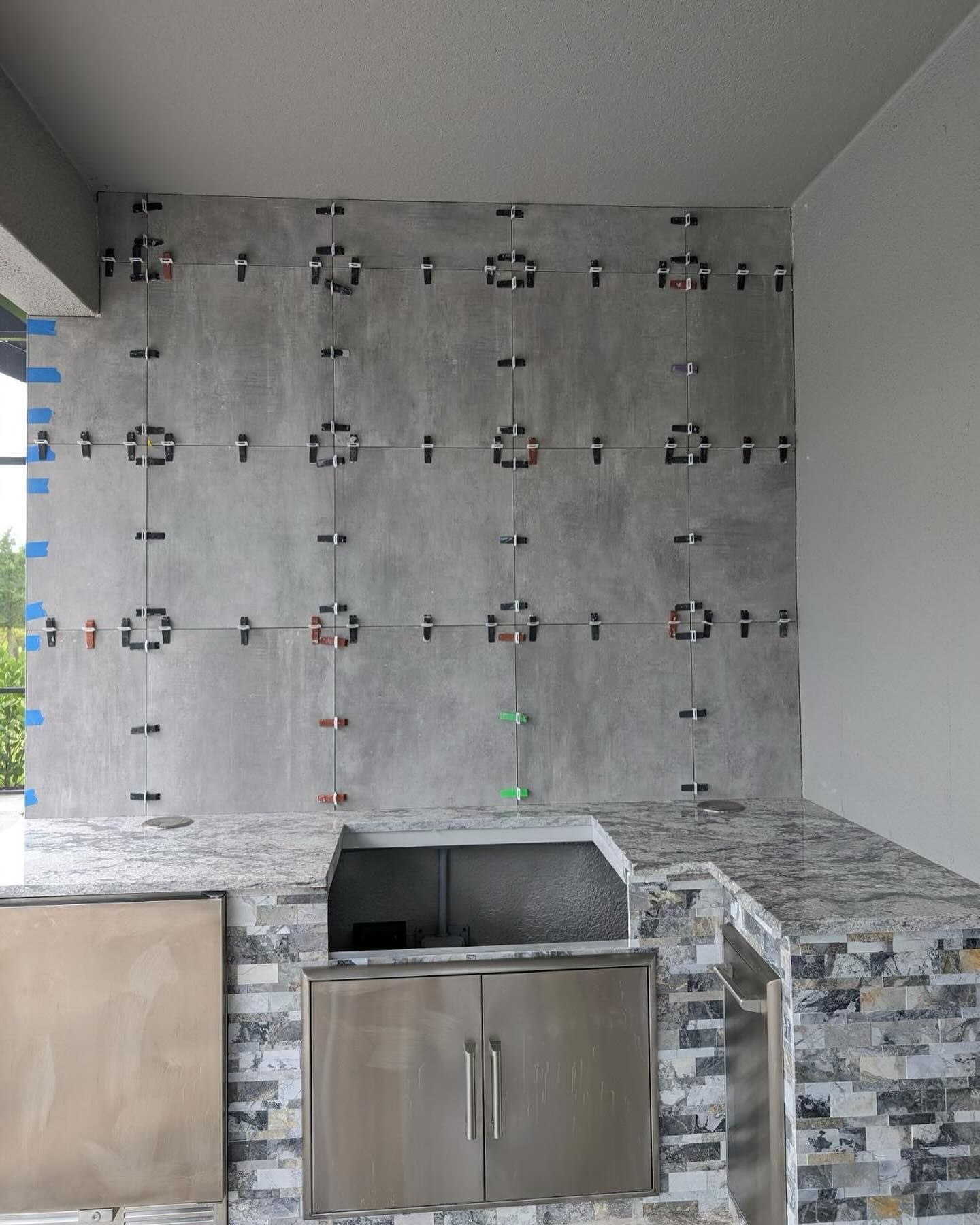 Outdoor kitchen under construction, gray tiled backsplash with spacers, stainless steel appliances, stone countertop.