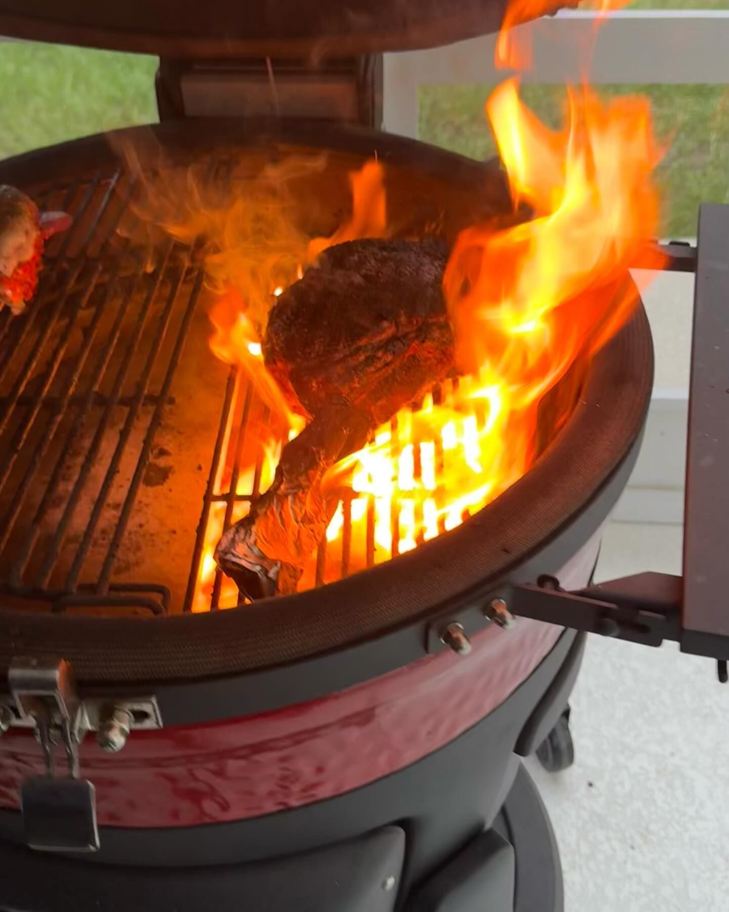 Red ceramic grill with flames engulfing a dark meat item on the grate. Outdoor setting.