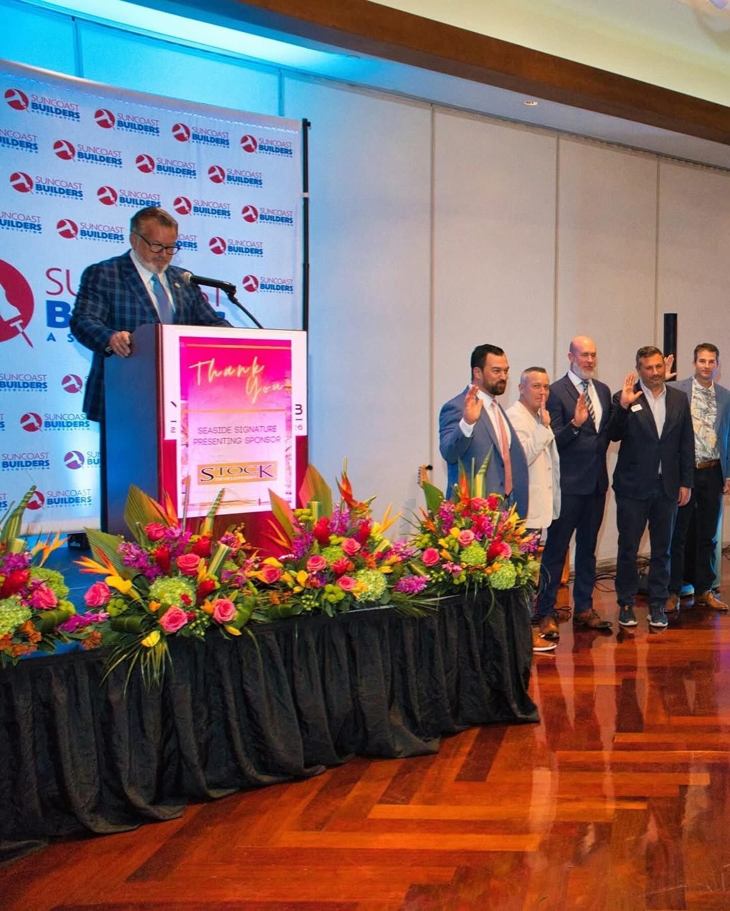 Man at podium speaks at an event, with people clapping. Colorful floral arrangements adorn the stage.