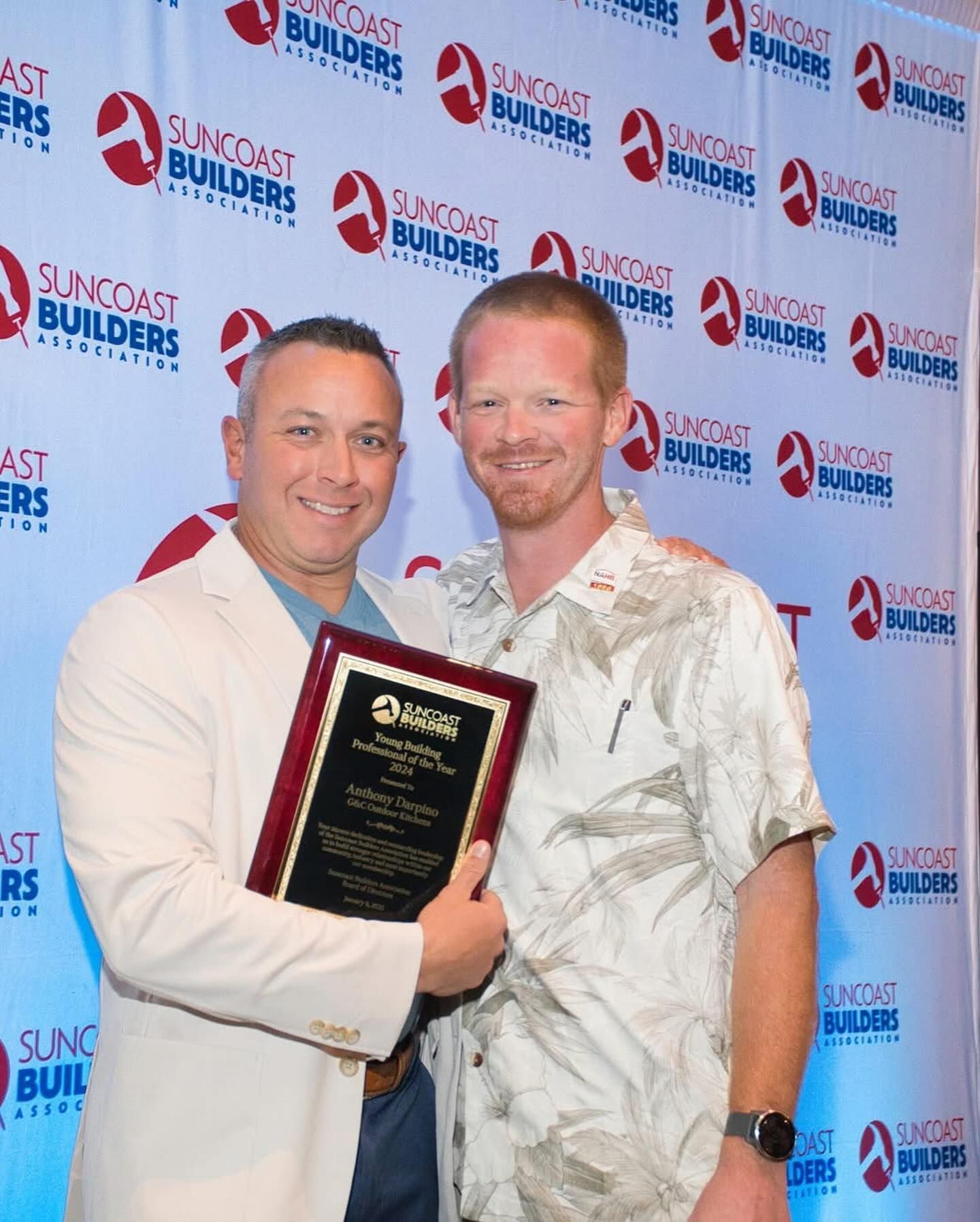 Two men holding an award plaque in front of a backdrop with logos. The man on the left is in a blazer. The other man is wearing a floral shirt.