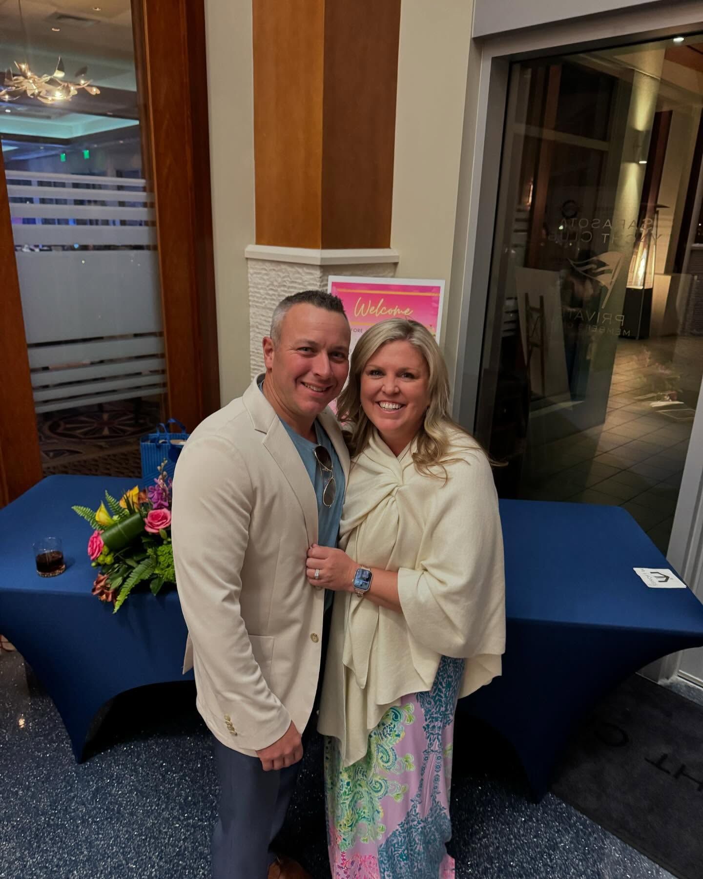 Couple smiling, embracing at an event; woman in a flowing dress and shawl, man in a blazer; navy table with flowers.