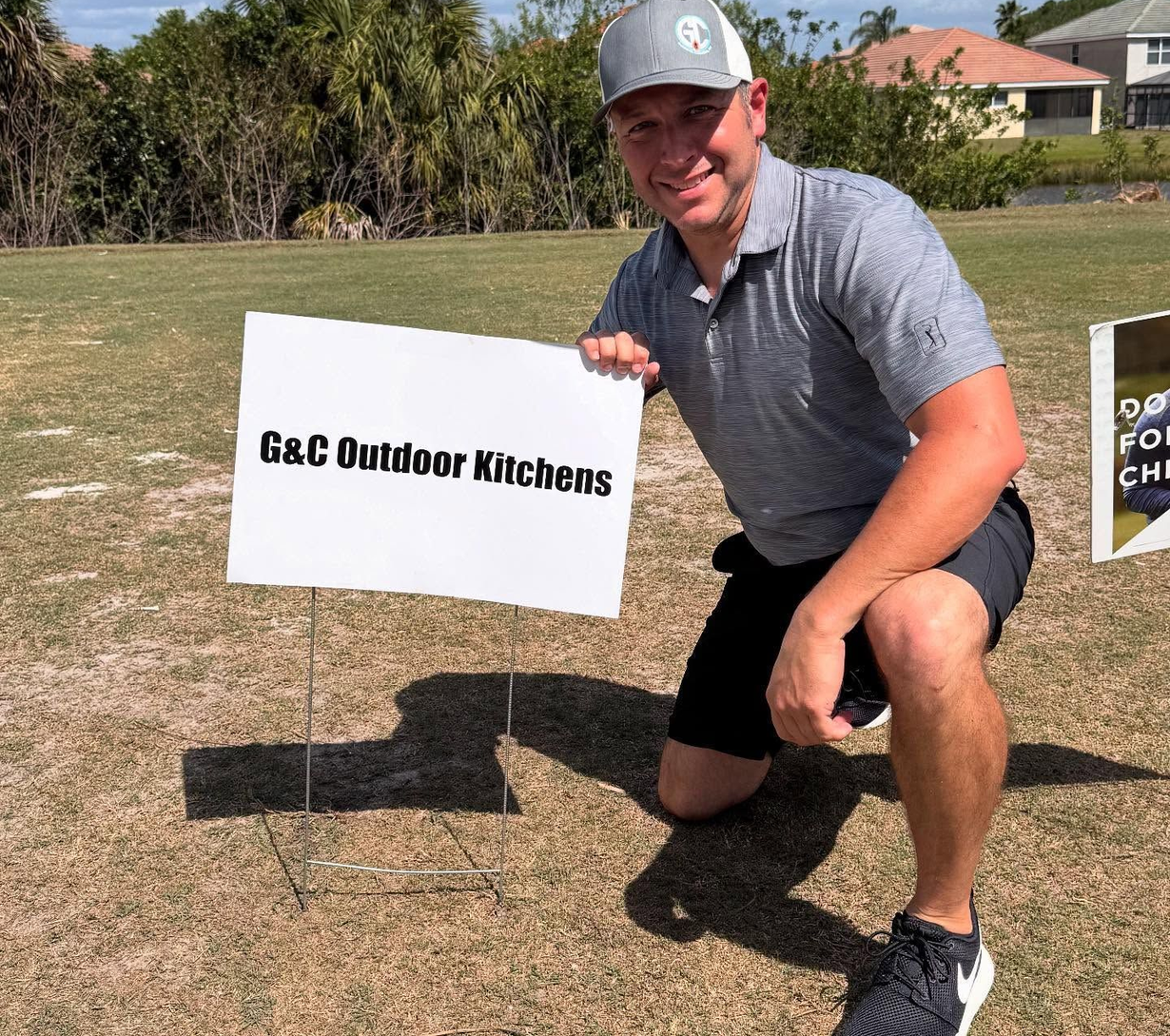A person kneeling in a grassy field holding a sign that reads G&C Outdoor Kitchens.