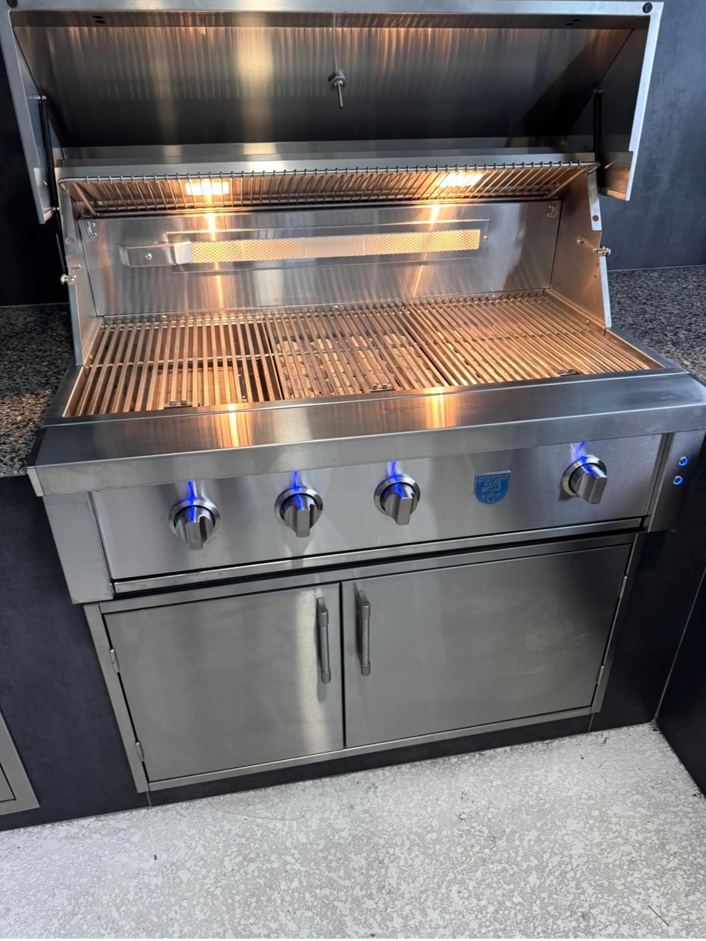 Stainless steel outdoor grill with open lid, lit burners, and storage cabinet.