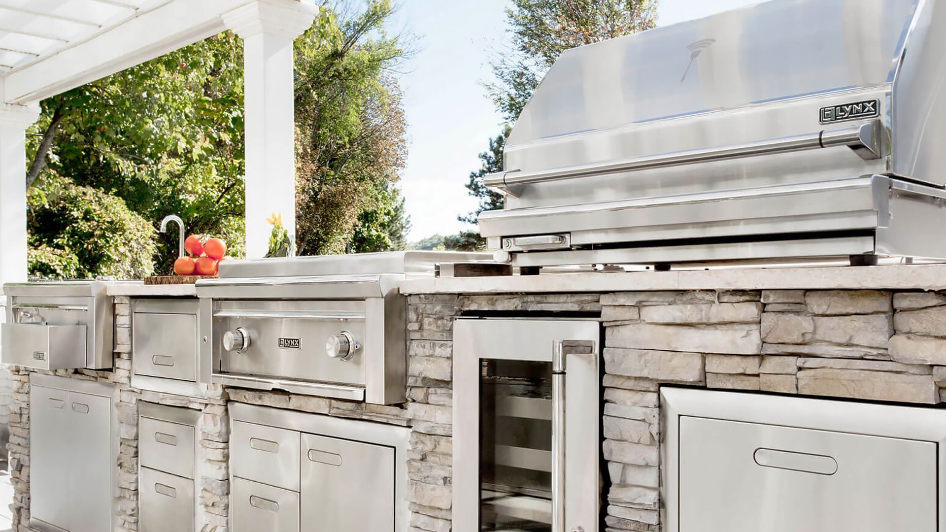 An outdoor kitchen island with a stone base, stainless steel grill, beverage cooler, and cabinets under a white pergola.