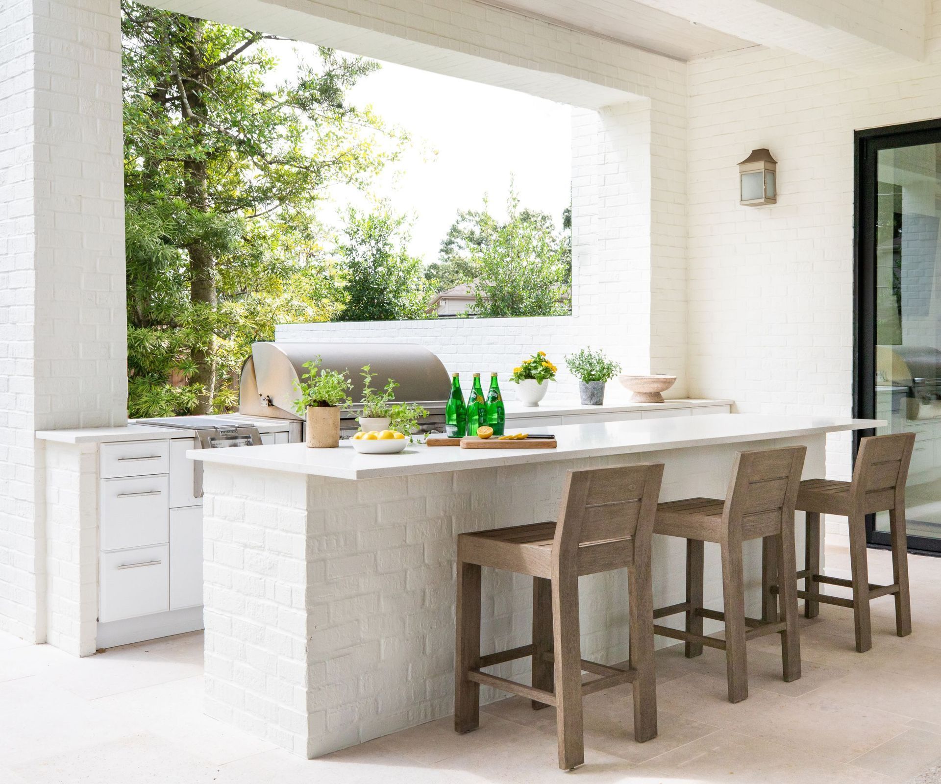 Bright outdoor kitchen with white counter, wooden stools, and potted plants under a covered patio