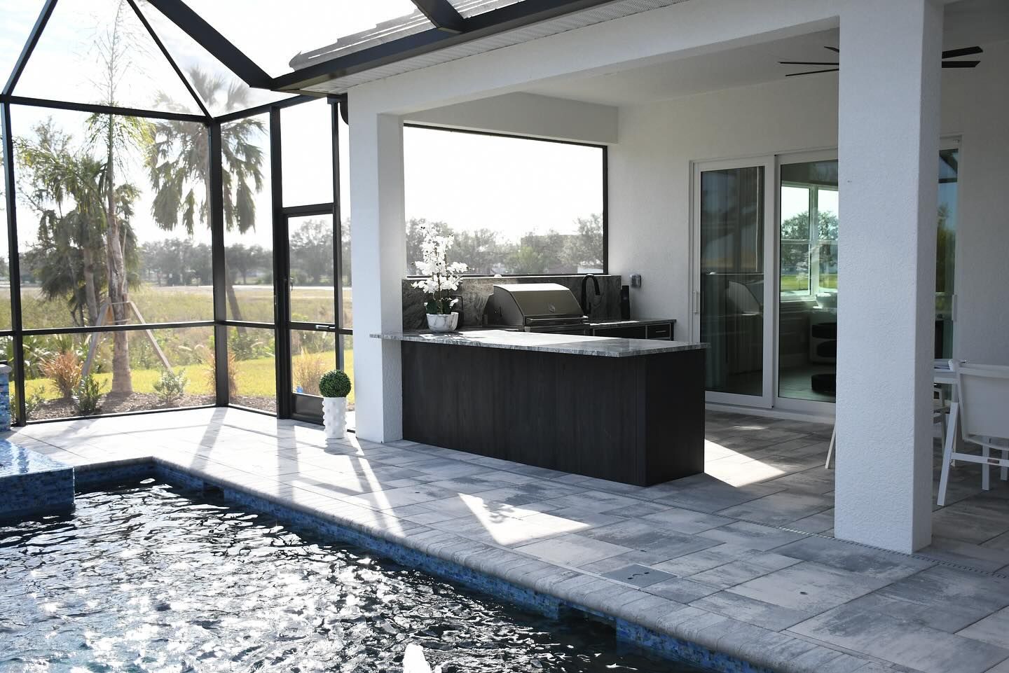 Modern patio with outdoor kitchen, lounge seating, and glass enclosure beside a pool.