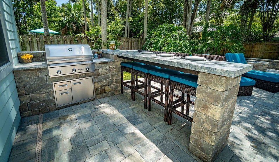 Outdoor stone patio with grill, bar counter, and stools beside a wooded backyard