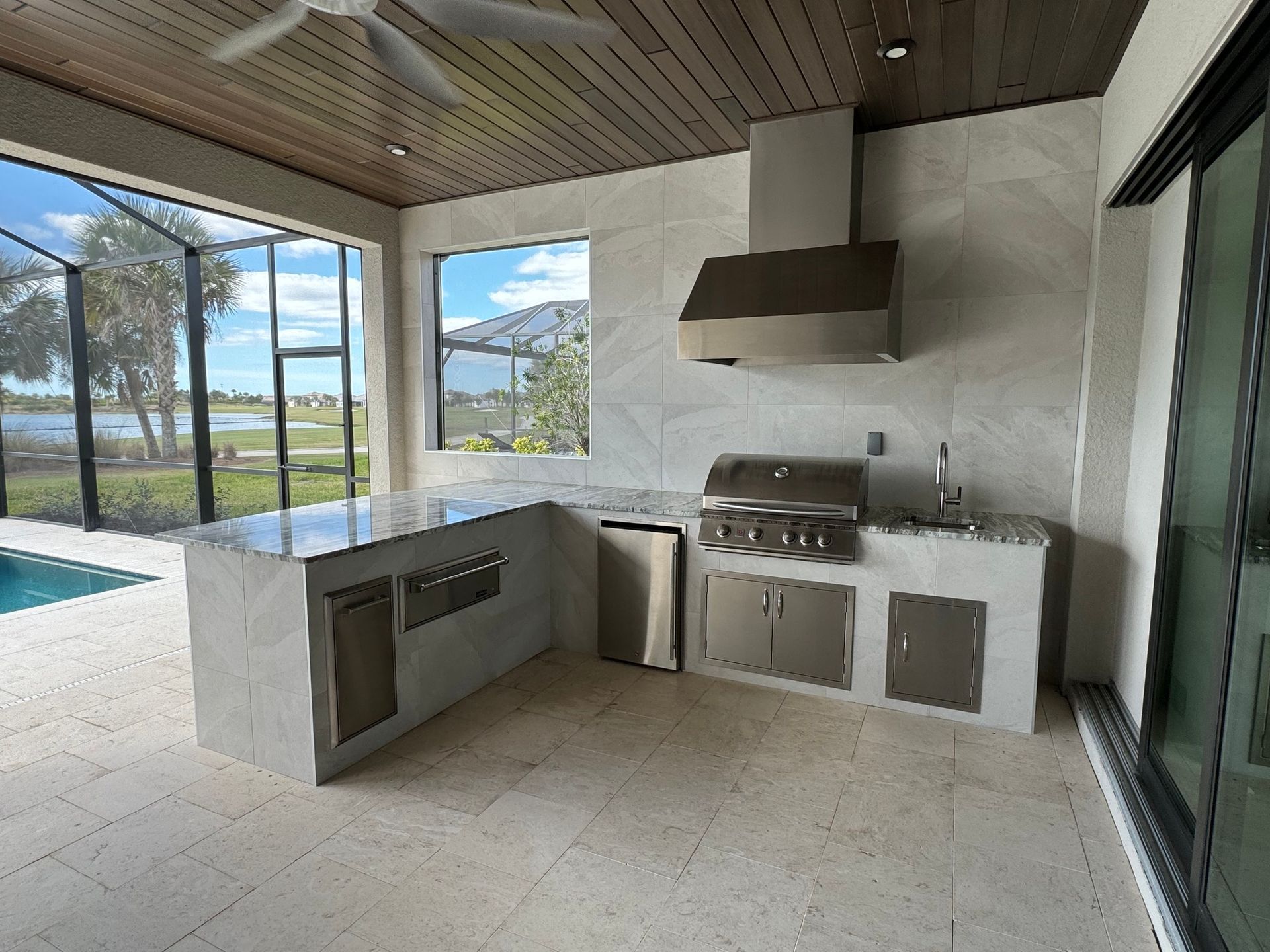 Outdoor patio kitchen with grill, sink, cabinets, and large windows overlooking water and trees