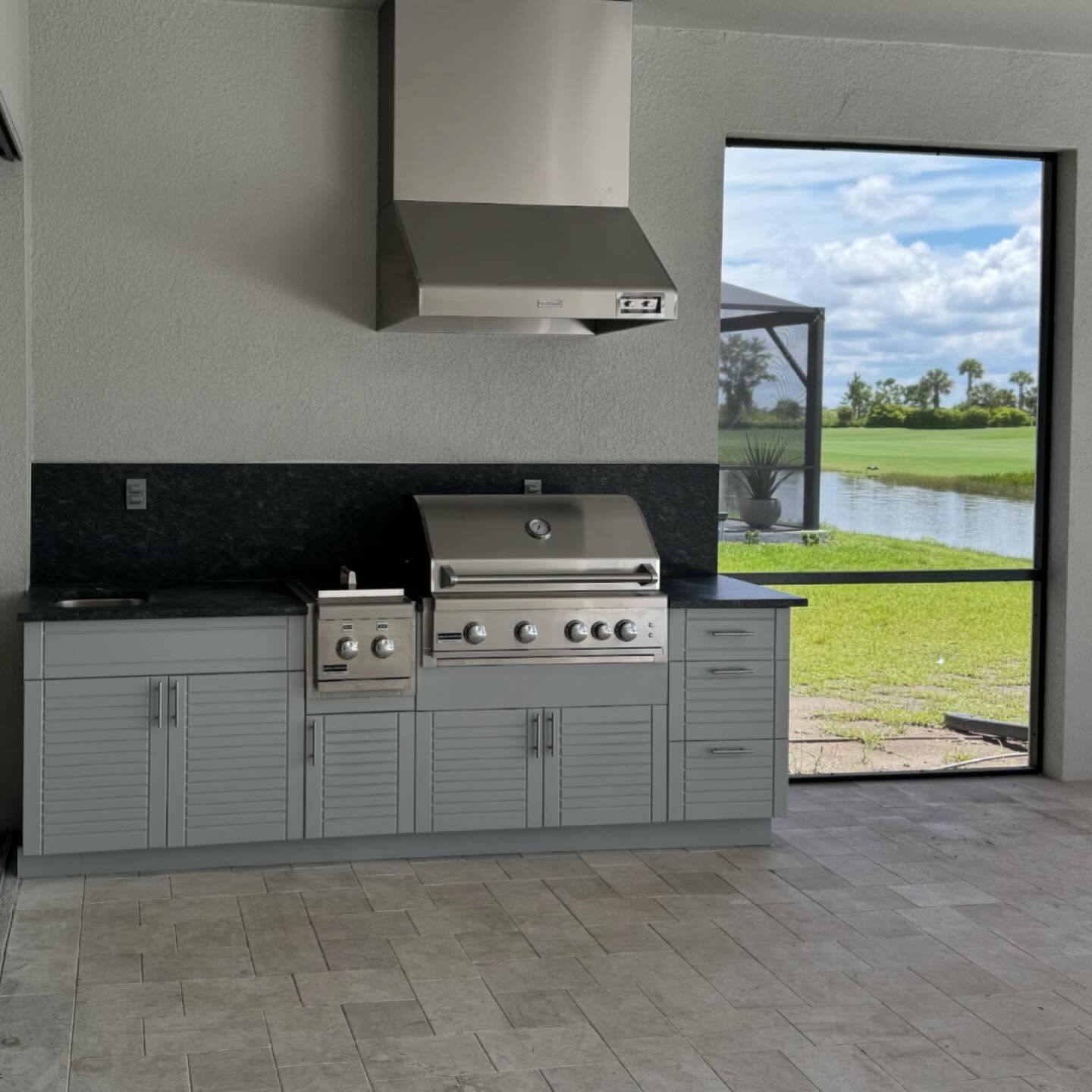 Outdoor kitchen with stainless steel grill, gray cabinets, and a black backsplash beside a patio and lawn