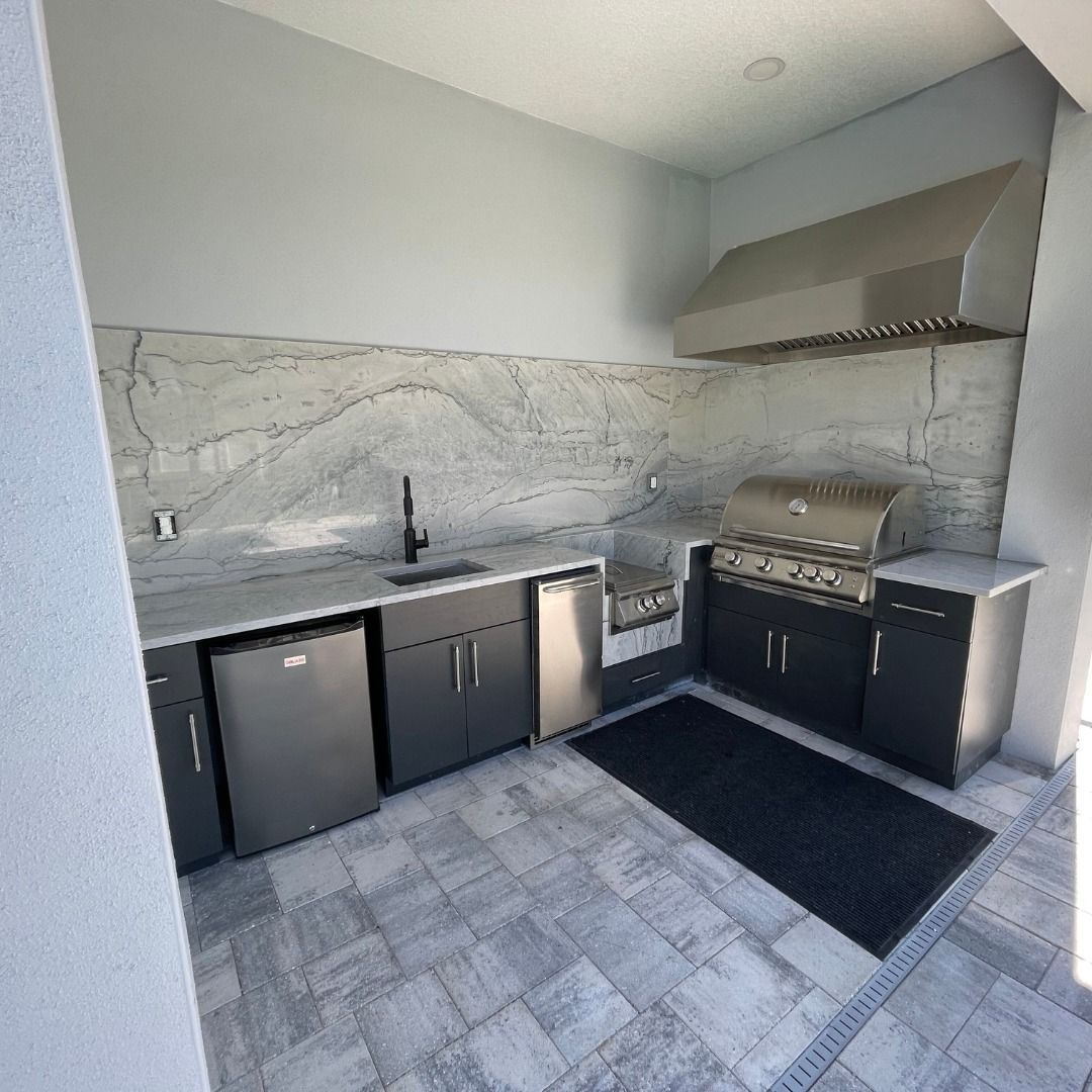 Modern outdoor kitchen with marble backsplash, black cabinets, sink, grill, and stainless mini fridge