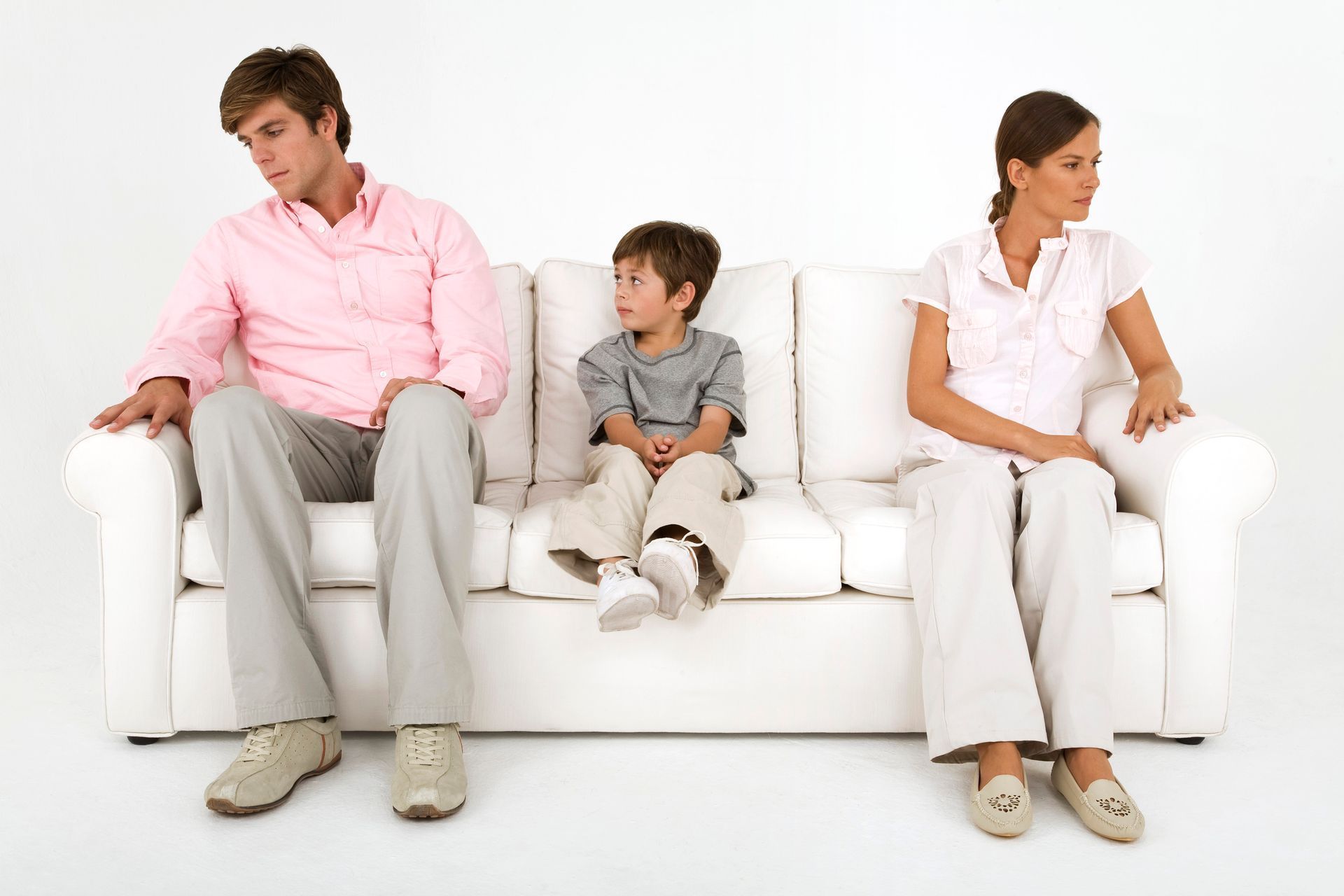 Three people sit apart on a white couch, looking away from each other in a bright living room.