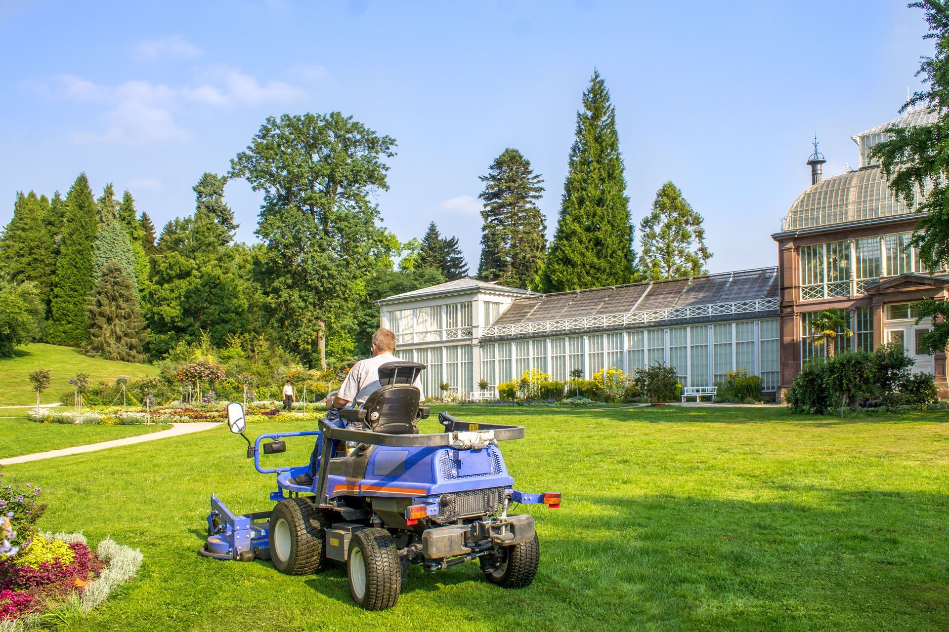 Person mowing a large lawn with a blue riding mower near a greenhouse and trees on a sunny day.