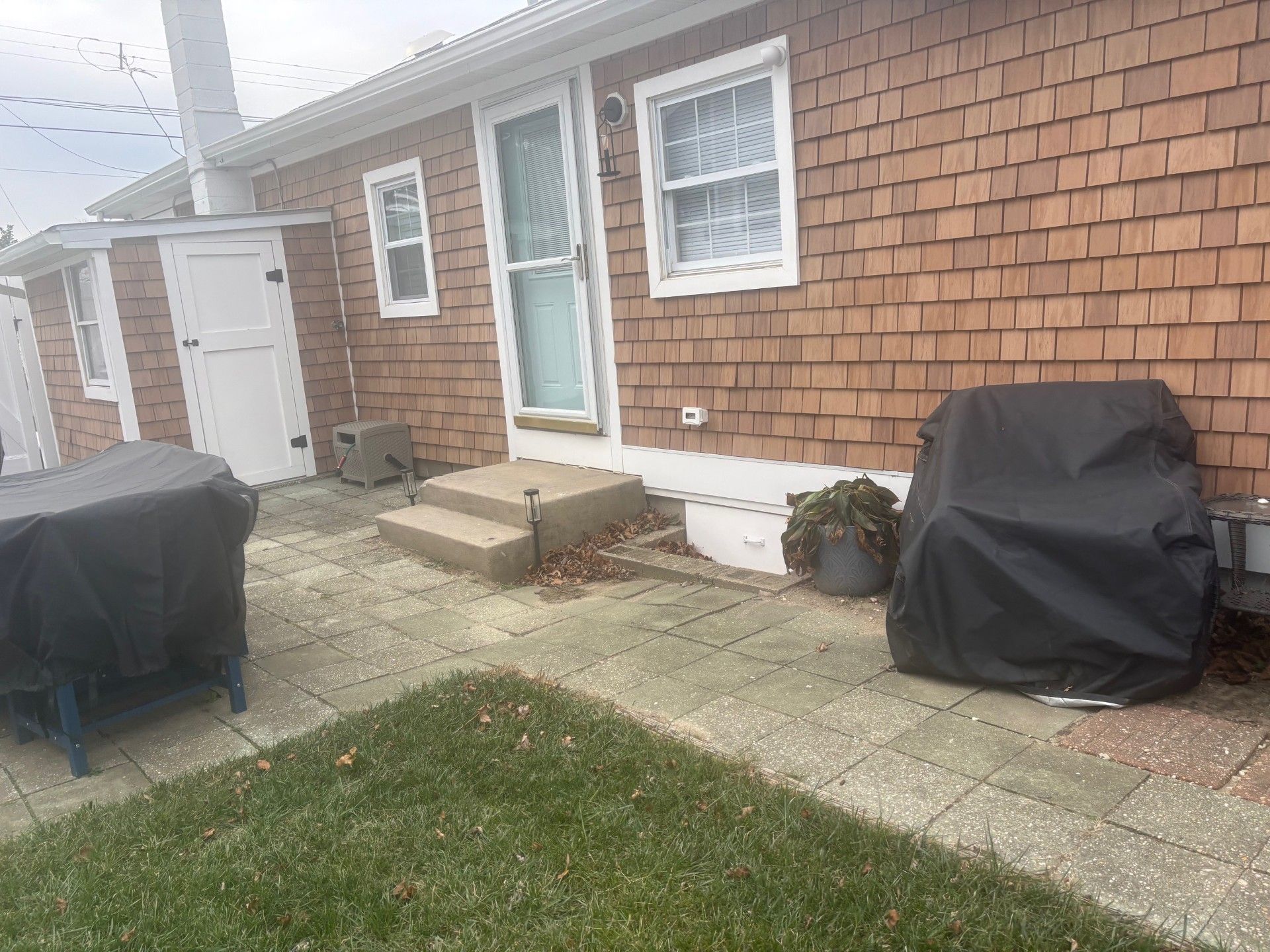 A patio outside a brick house with cedar shake siding, two covered grills, a small staircase, and a doorway.