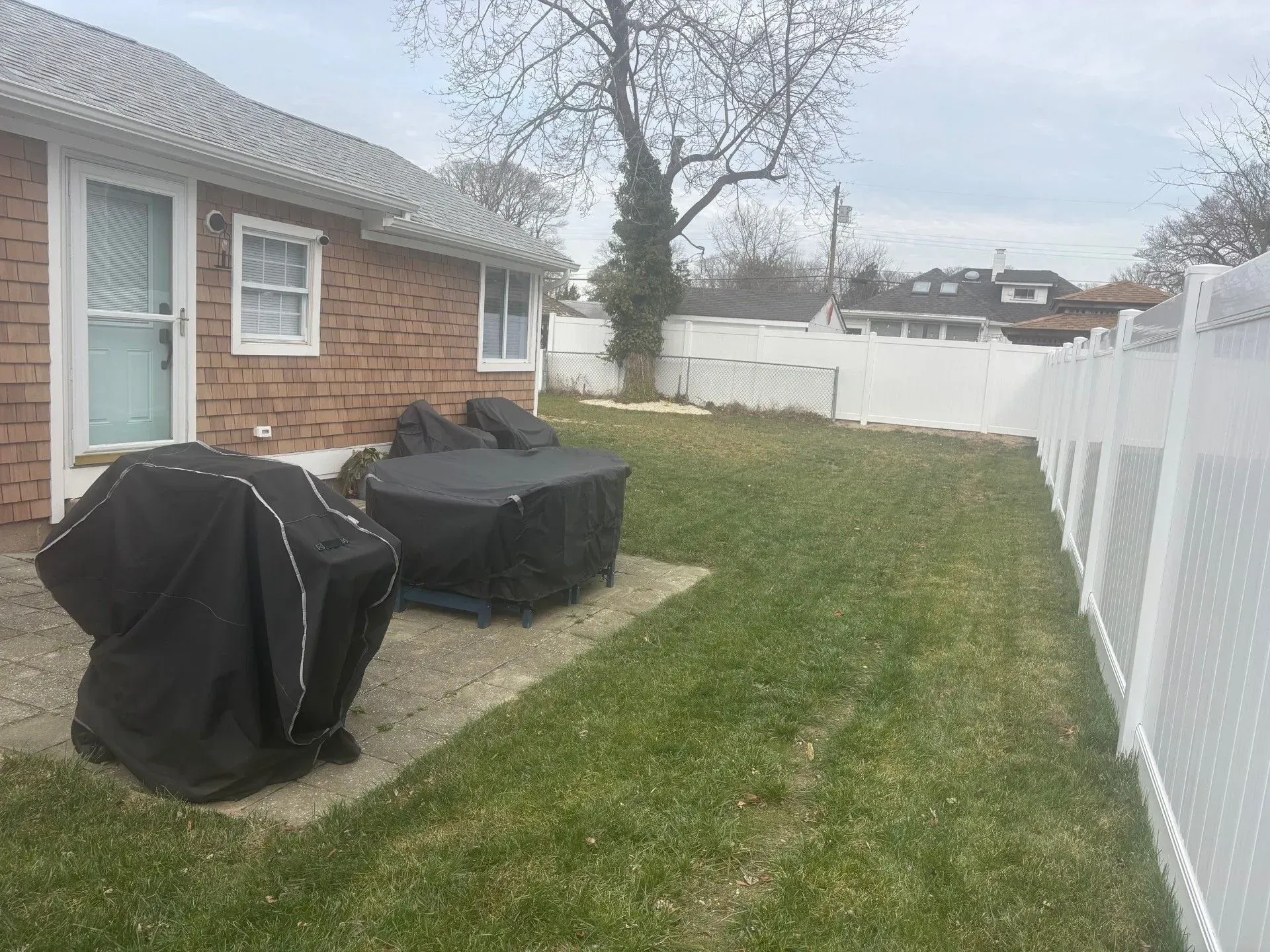 A backyard with a patio containing a covered grill and table, next to a brick house and a white vinyl privacy fence.