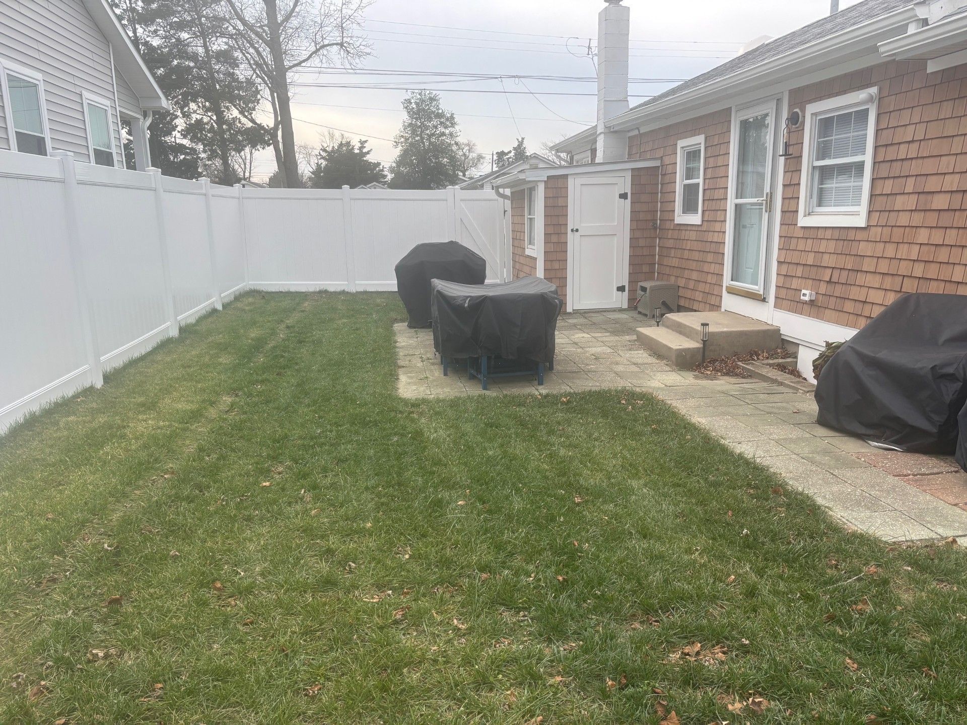 A grassy backyard featuring a concrete patio with outdoor furniture covered by black tarps, enclosed by white fencing.