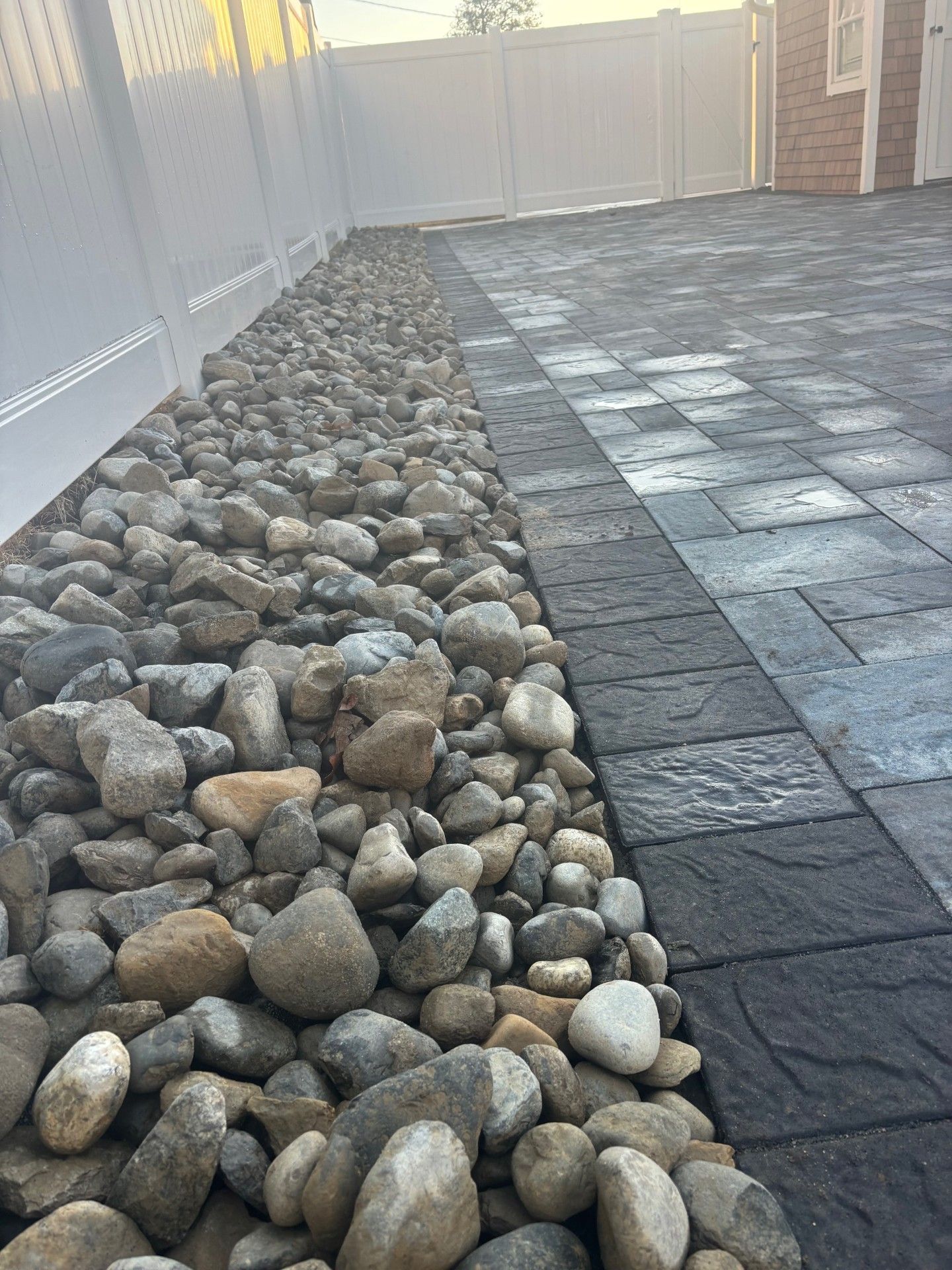 A gray stone pathway bordered by a strip of river rocks next to a white fence in a backyard.