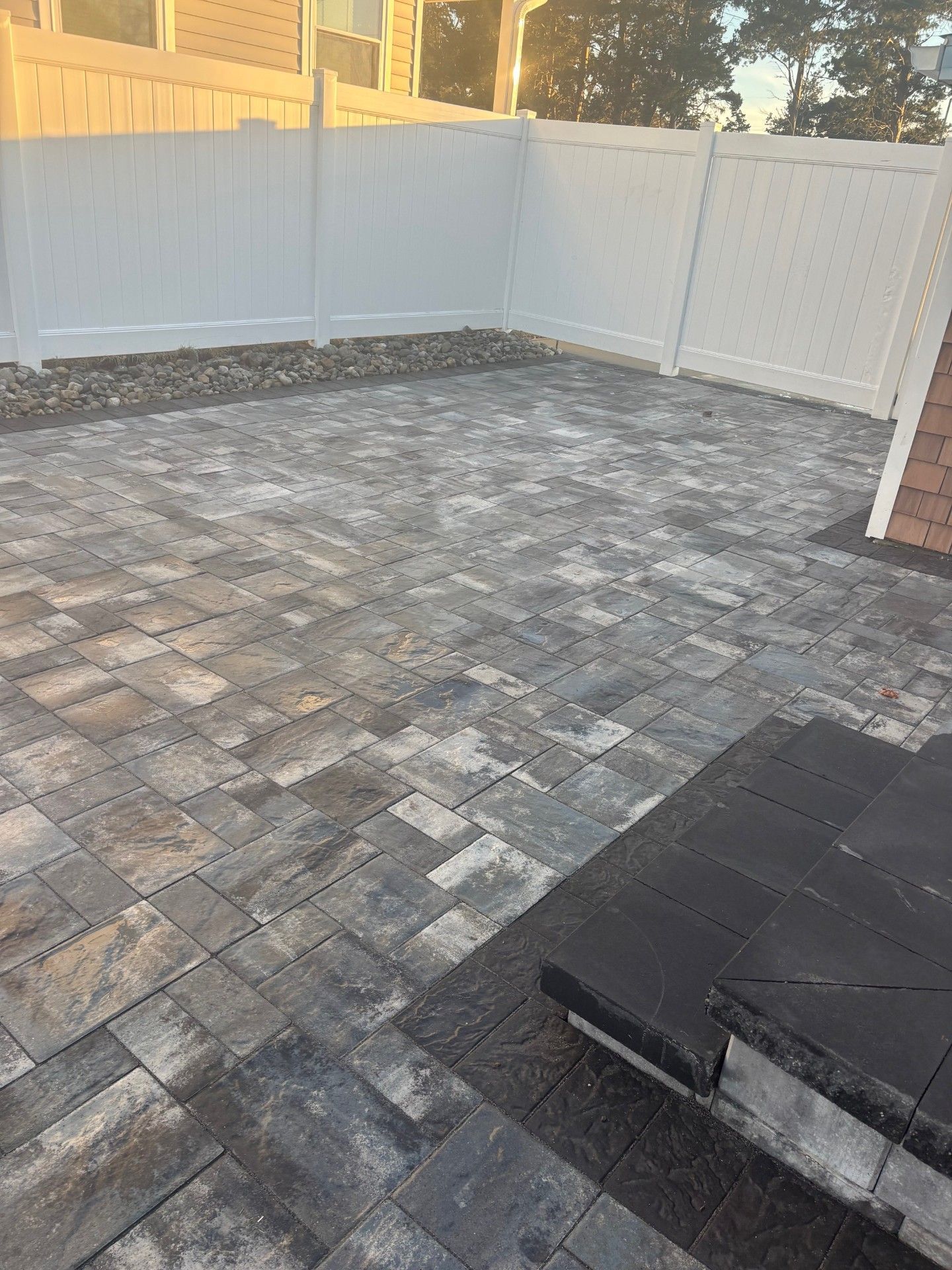 A gray stone patio with a patterned design, bordered by a white fence and a set of dark stone steps leading to a building.