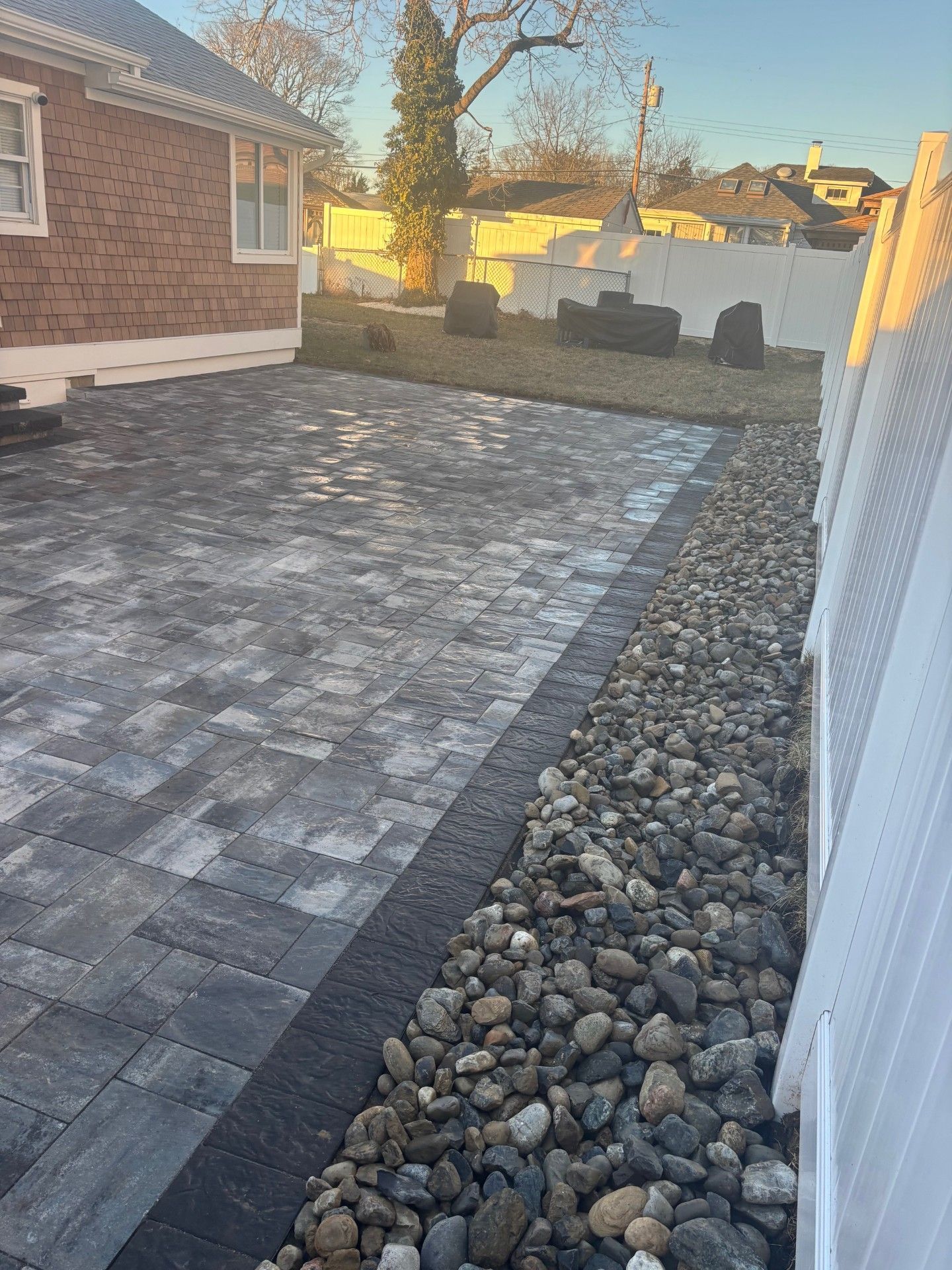 A stone patio with a dark border next to a gravel strip along a white fence, with a brick house visible in the background.