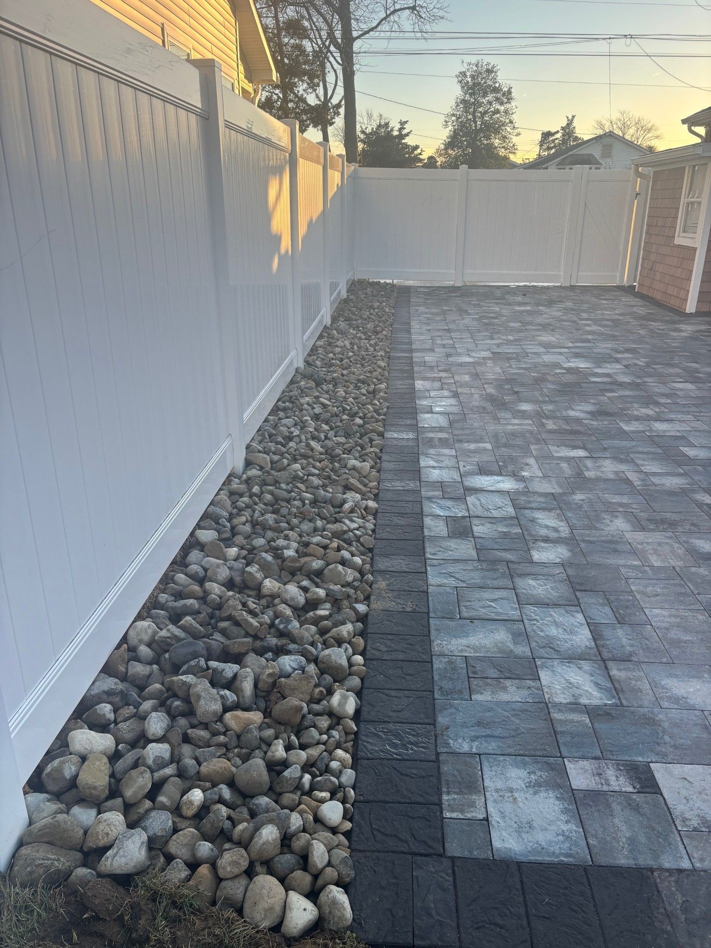A white vinyl fence borders a gravel garden bed next to a paved stone patio.