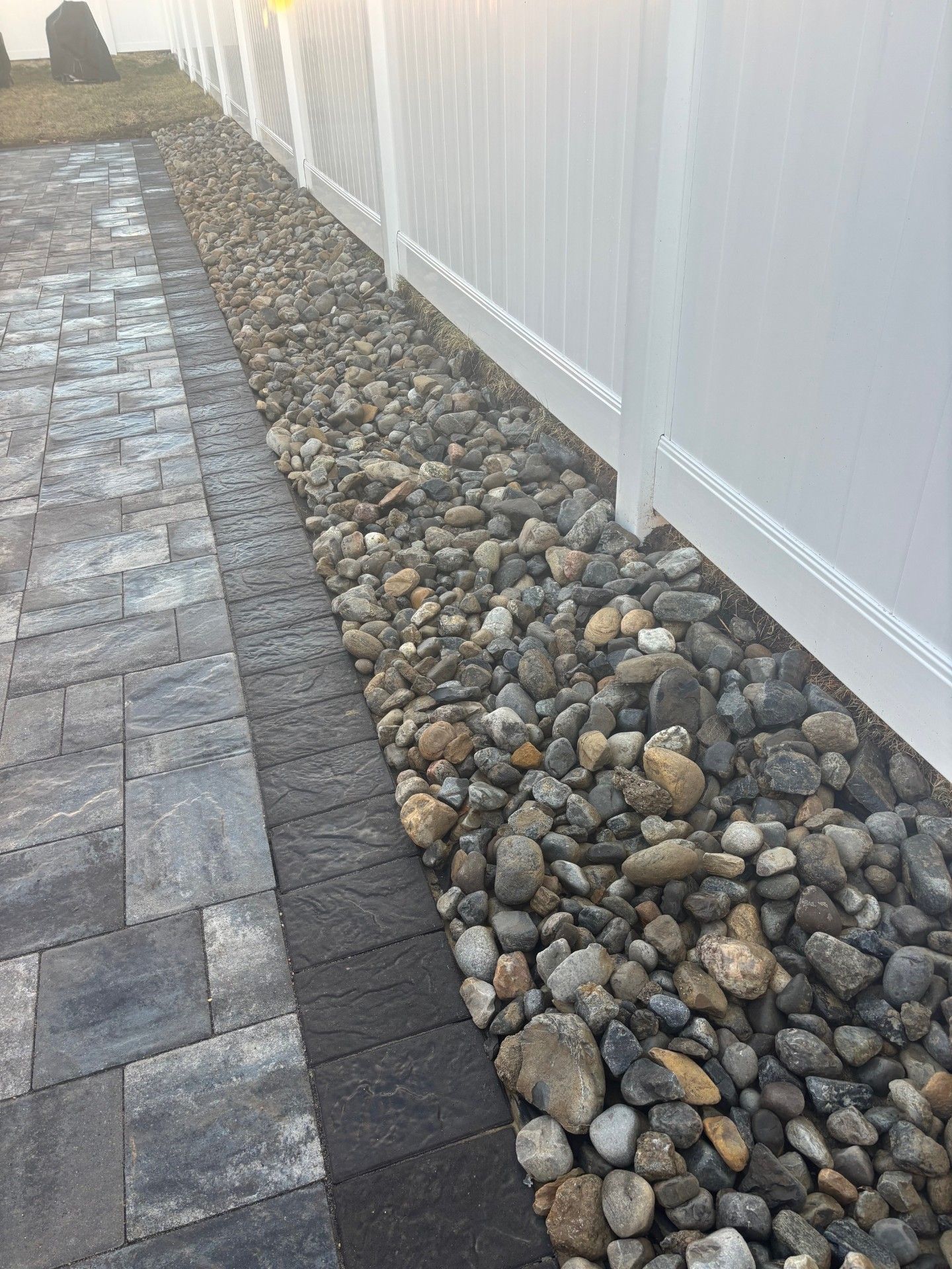 A row of multi-colored landscaping river stones beside a white vinyl fence and gray paver walkway.