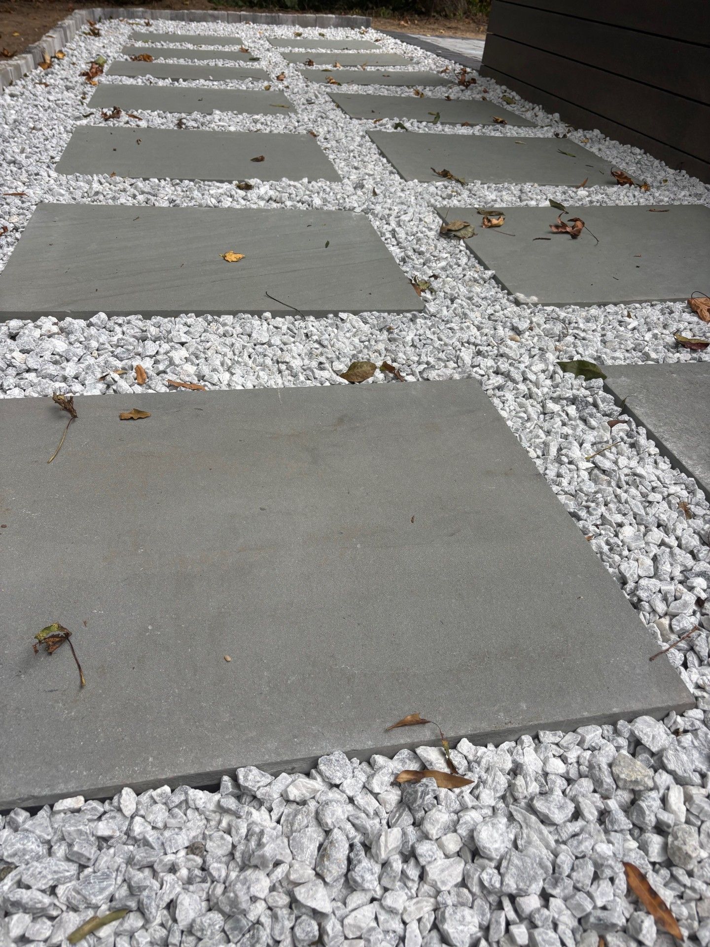 A path of large, grey square concrete pavers set in a bed of white decorative gravel along a dark wall.