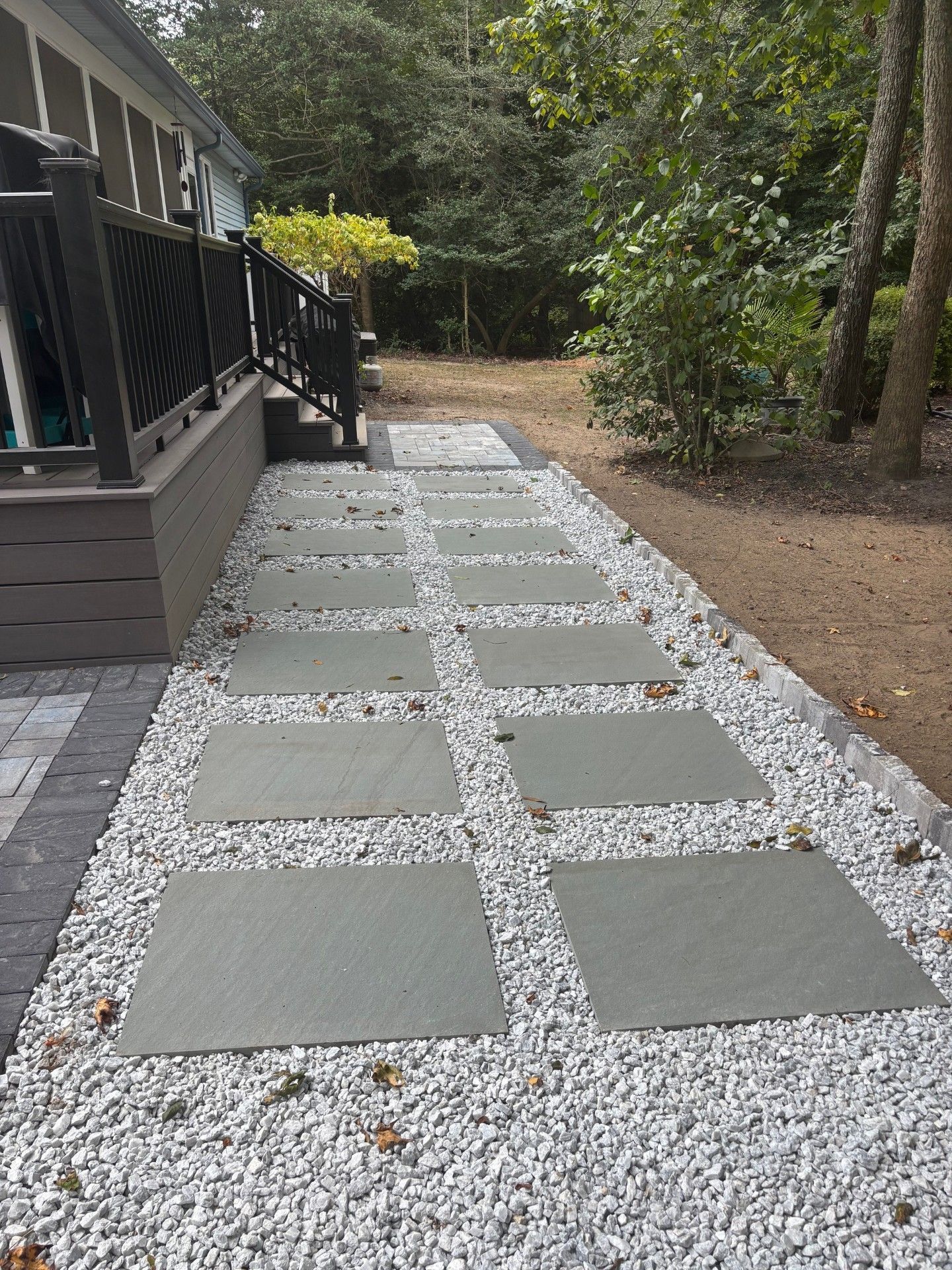 A stone paver walkway with white gravel set beside a wooden deck and trees in a backyard setting.