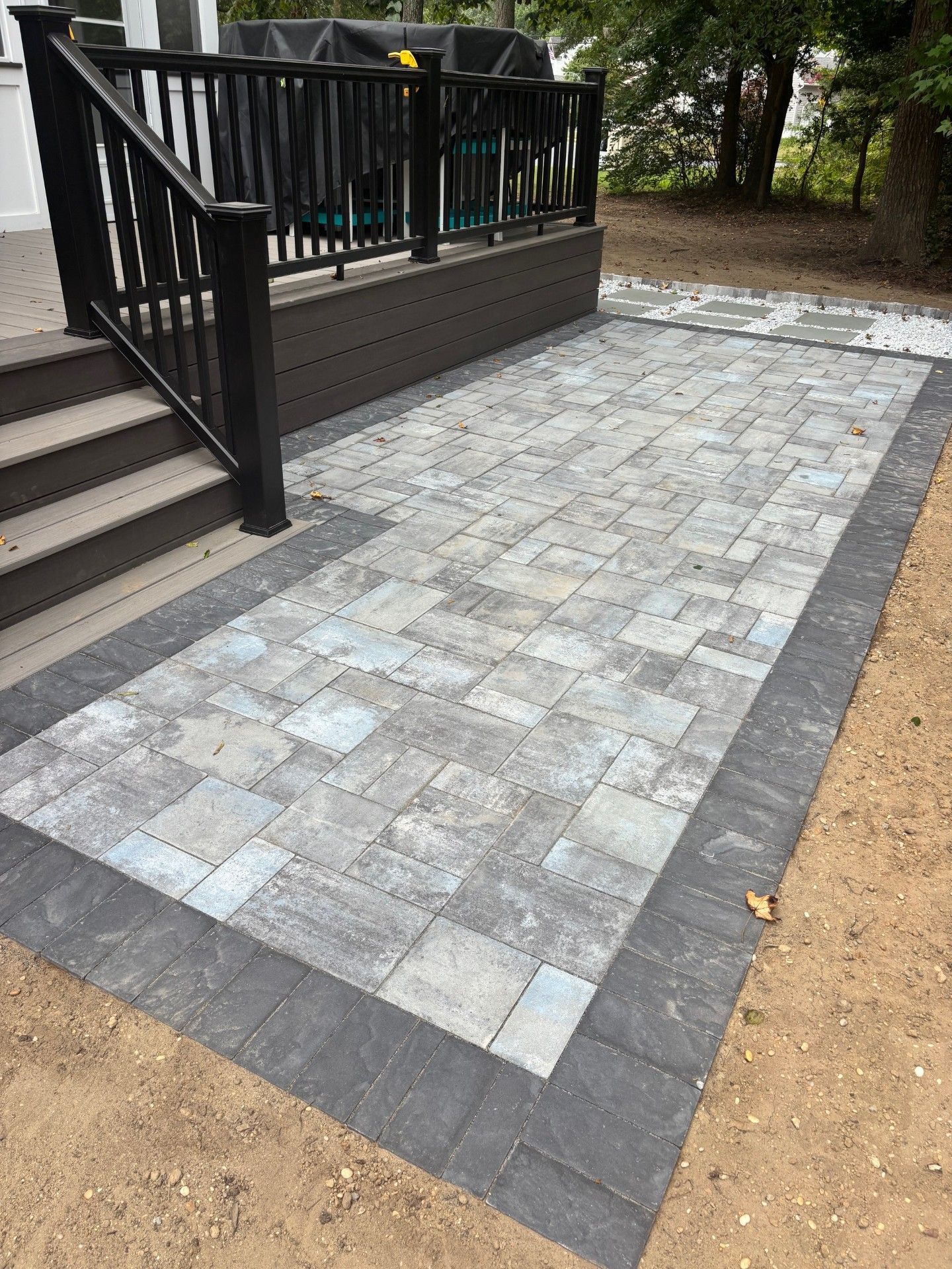 A paved patio with grey patterned pavers and a dark border, situated beside a composite deck with black railings.