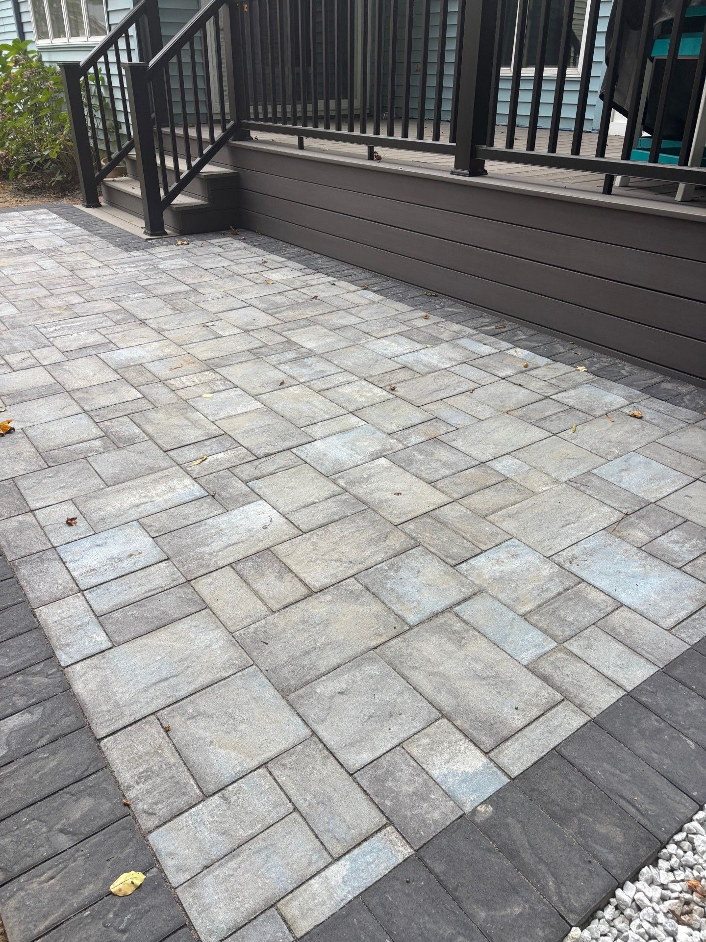 A patio paved with rectangular gray stones, bordered by dark bricks, next to a raised wooden deck with stairs.