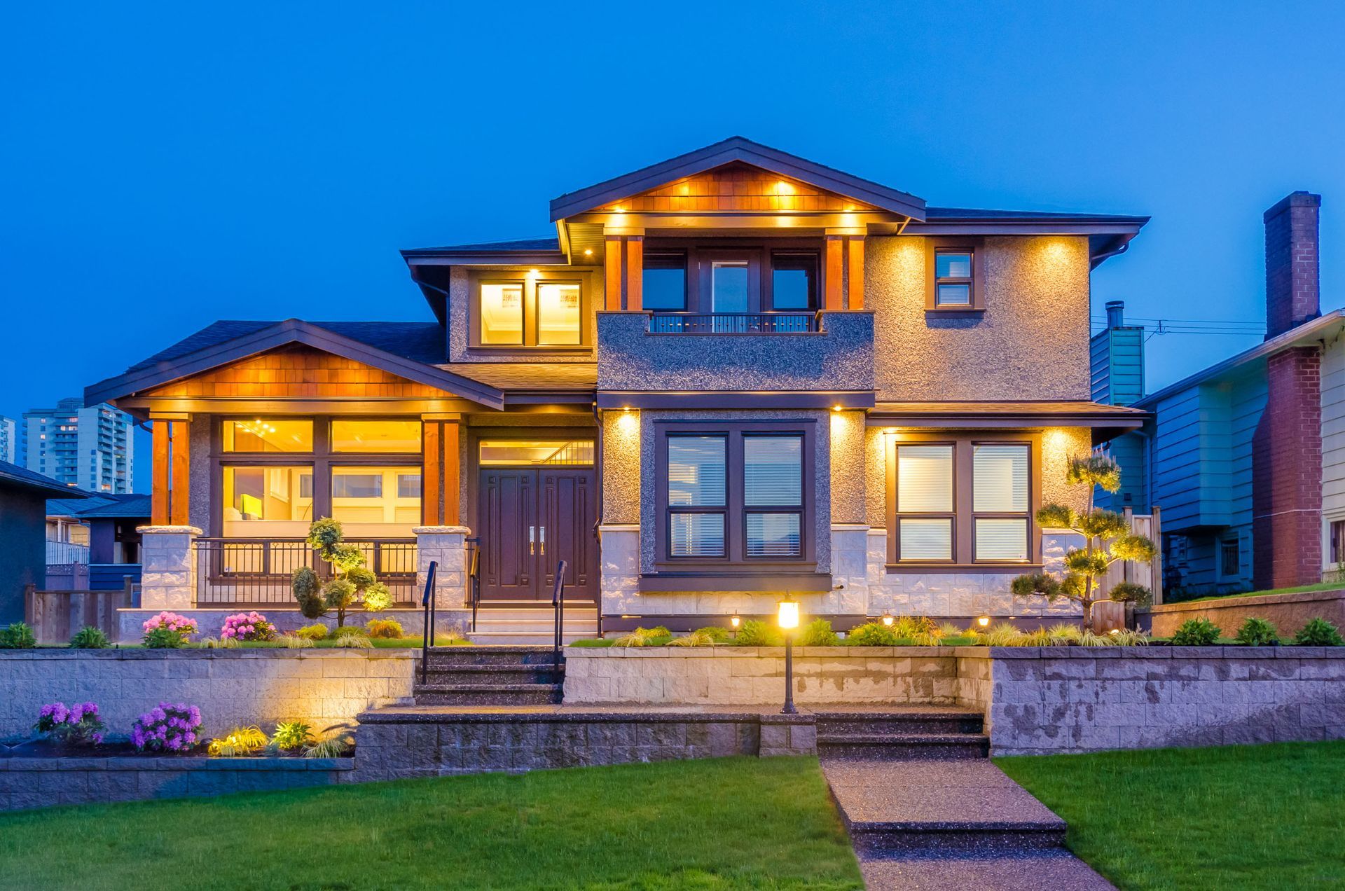 Lit up multi-story house at dusk with stone facade, dark doors, and manicured landscaping.