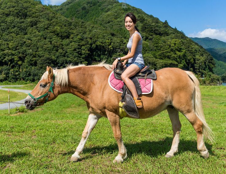 Woman smiling, riding a light brown horse in a grassy field near mountains.