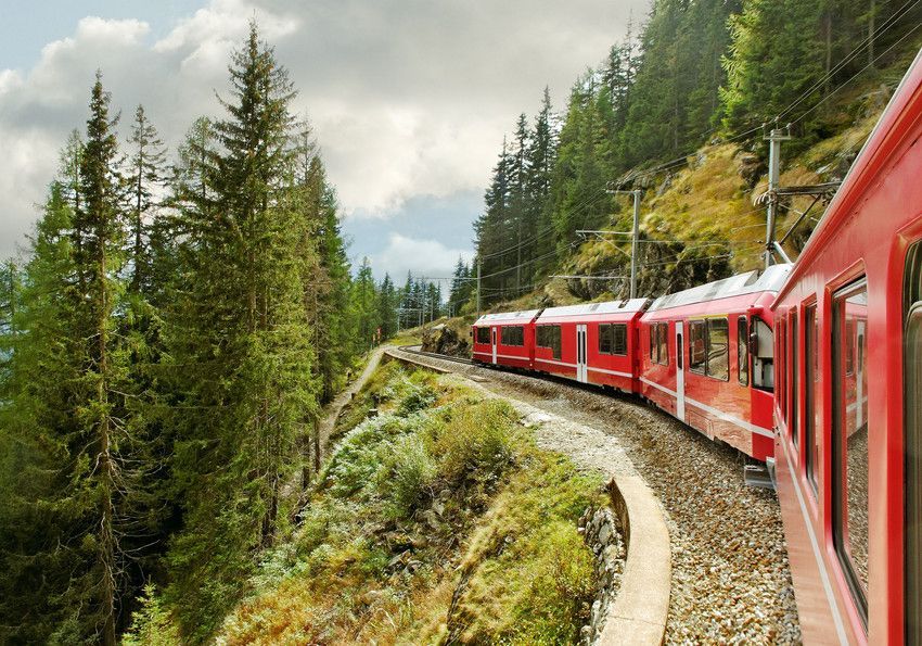 Red train winding through a green, mountainous forest under a cloudy sky.