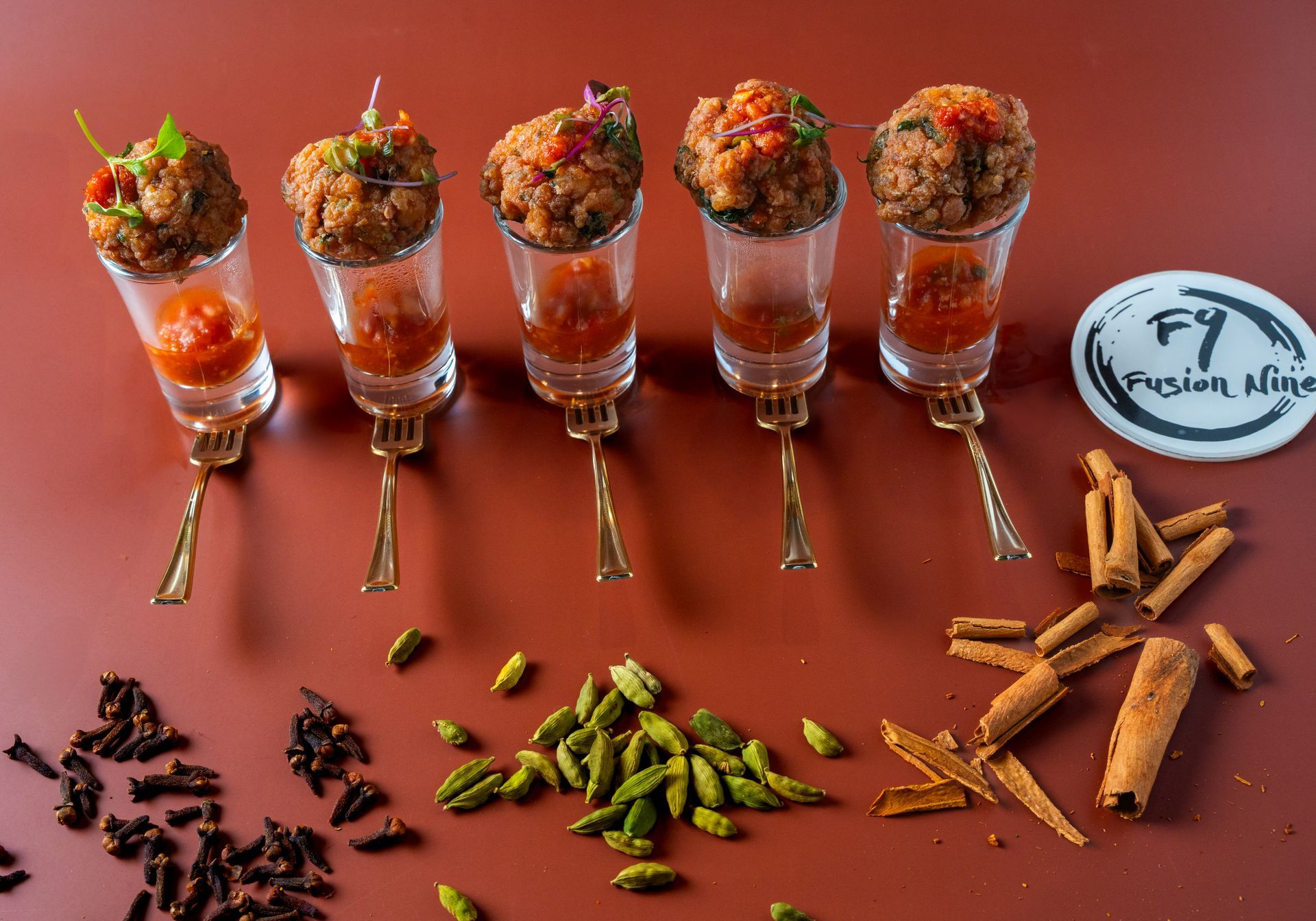 Appetizer meatballs in shot glasses with sauce, on a reddish-brown surface with spices and the