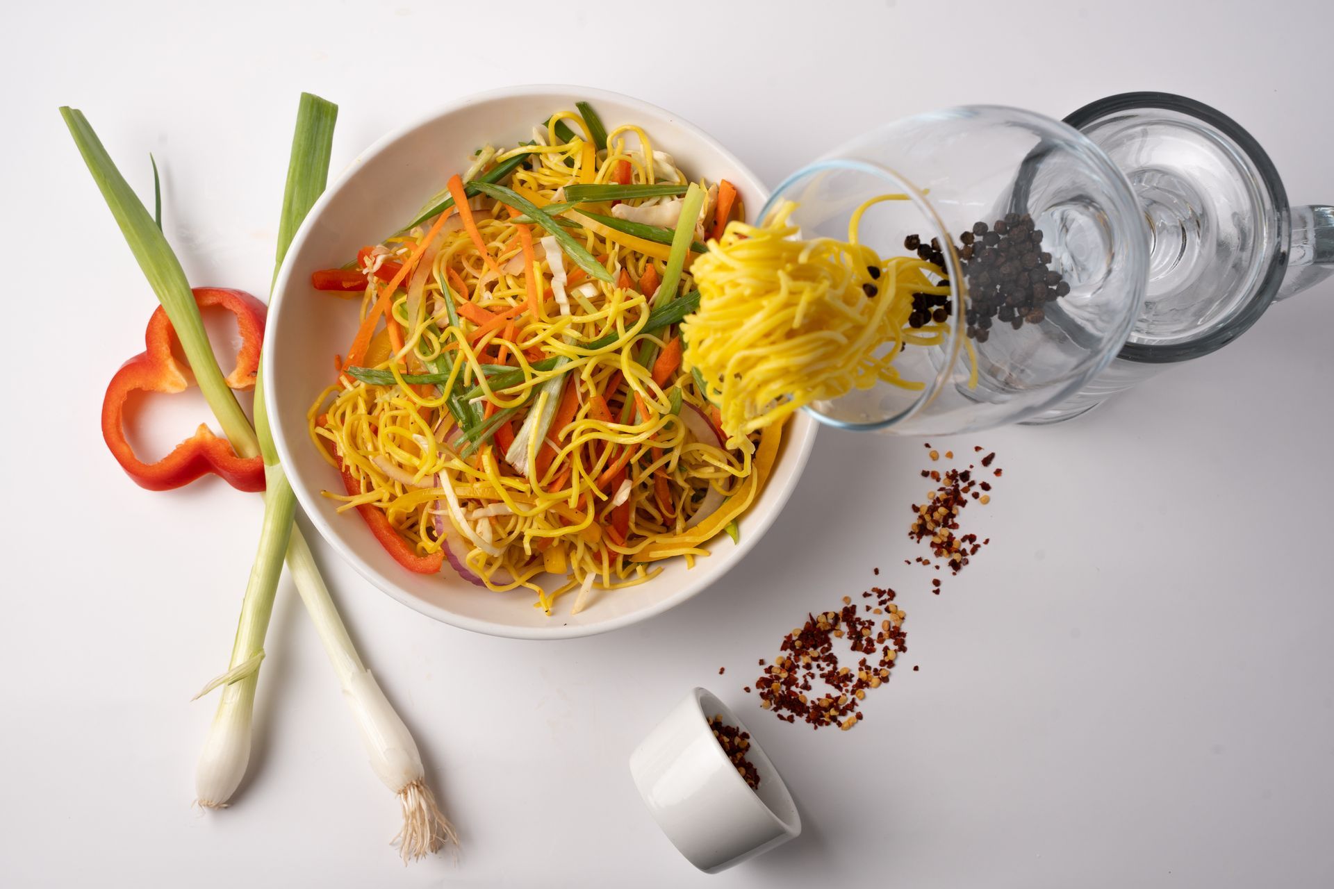 A bowl of yellow noodles with vegetables and a glass spilling seasoning on a white surface.