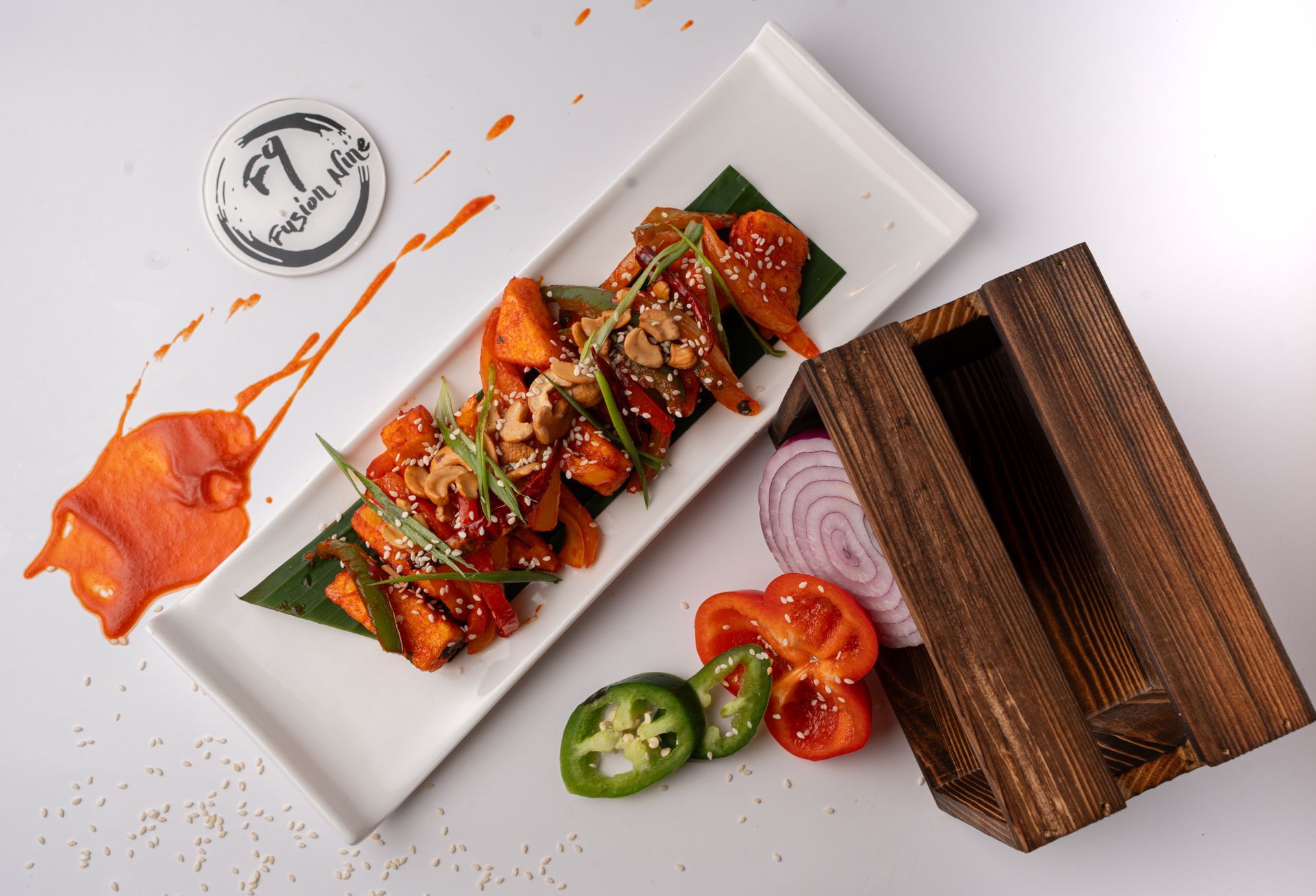 A rectangular plate of food with orange sauce and toppings. Garnishes include red onion, peppers, and a wooden box.