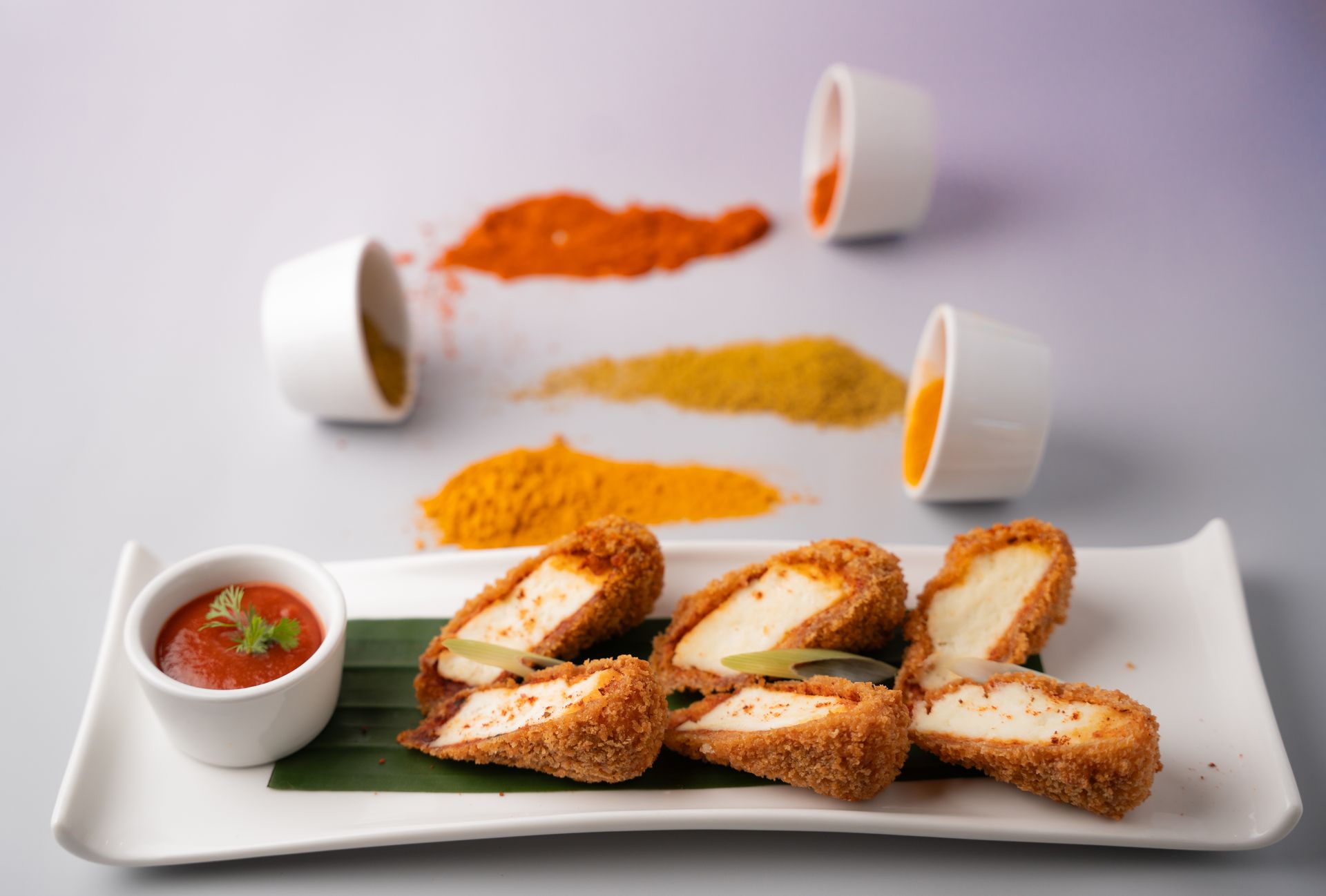 Breaded paneer cheese appetizer with dipping sauce, arranged with spilled spices and small bowls on a white plate.