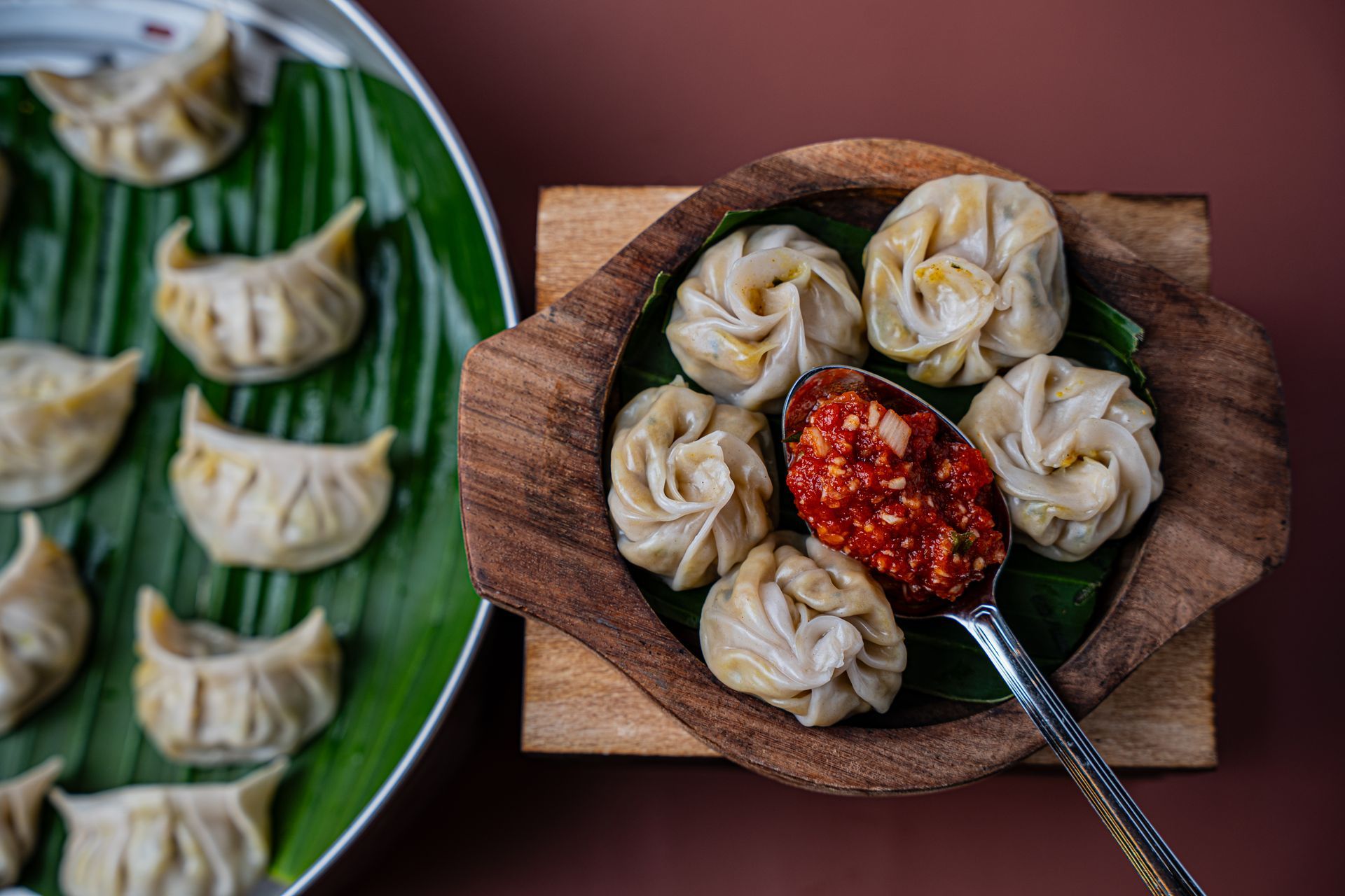 Steamed momos and spicy red chutney in a wooden bowl, served on a banana leaf. Momos also presented on a tray.