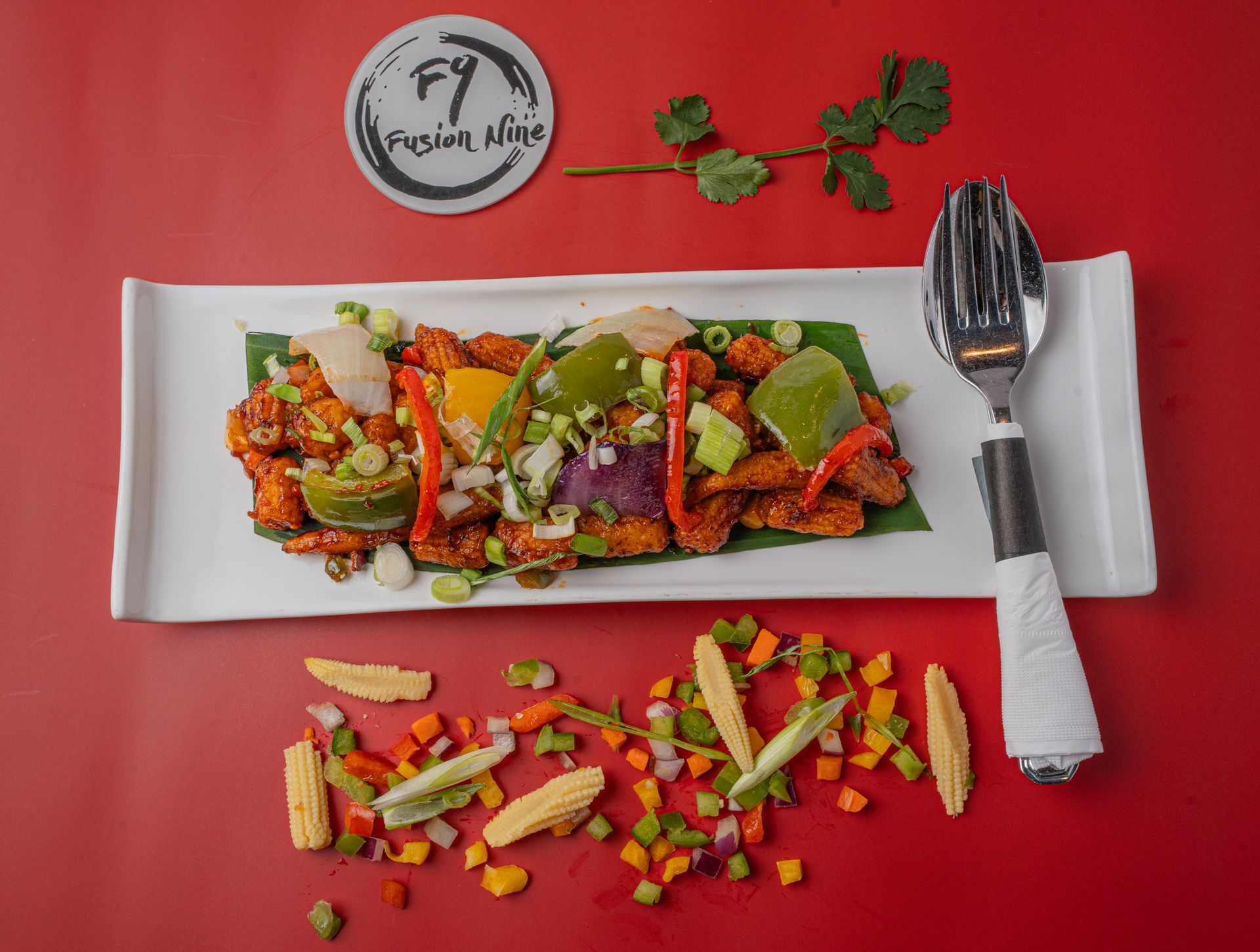 Chili chicken dish on a white platter with vegetables and a spoon. Garnished with herbs on a red surface.