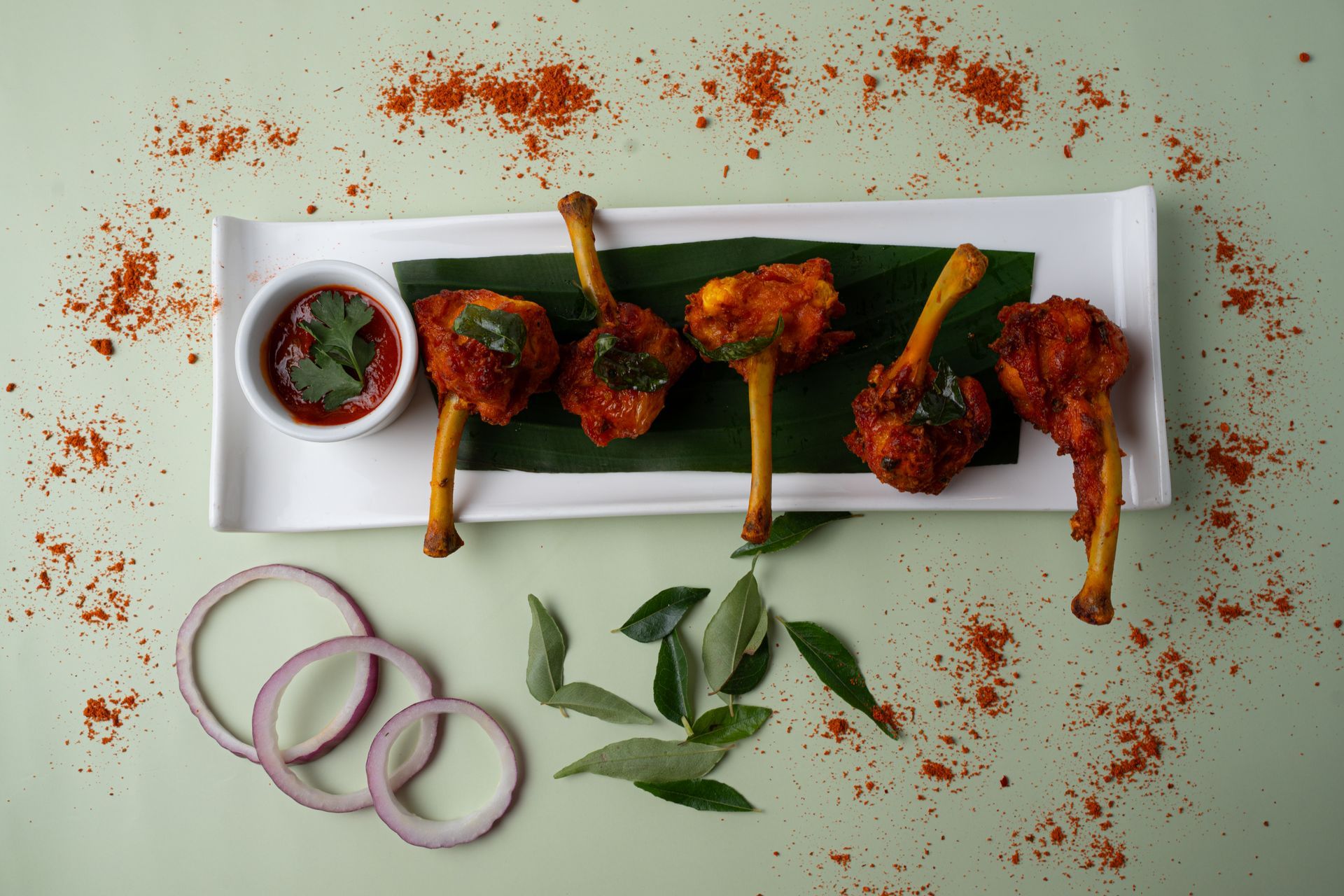 Fried chicken lollipops with a red sauce on a white platter with onion rings and scattered spices.
