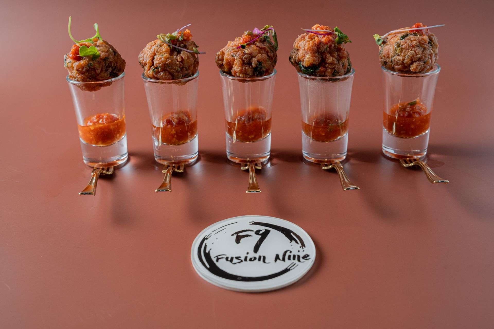 Five fried food balls in shot glasses with sauce, arranged on a brown surface with an FY Fusion Nine logo.