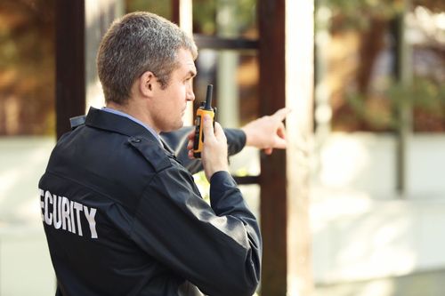 Security guard in dark uniform speaking into a radio and pointing outdoors.