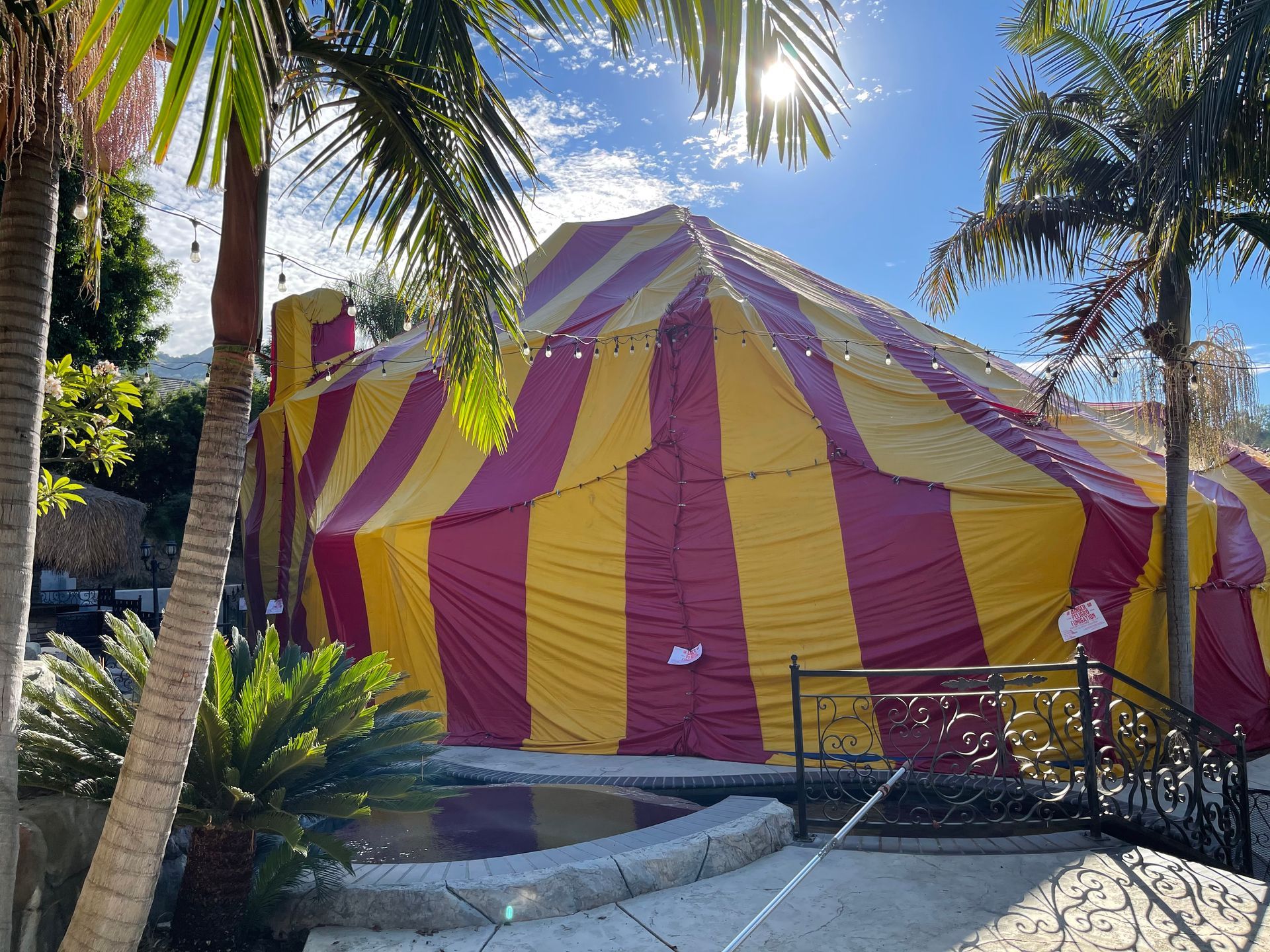 House tented with red and yellow striped tarp, surrounded by palm trees and greenery on a sunny day.