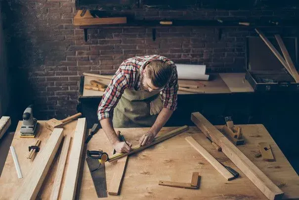 A woodworker wearing a plaid shirt and apron measures a piece of timber on a cluttered workshop workbench.
