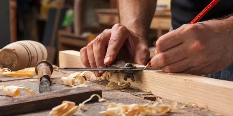 A person uses a marking gauge and a red pencil to draw a precise line on a wooden plank in a workshop setting.