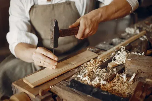 A person in an apron uses a hammer to nail a piece of light-colored wood on a workbench covered in wood shavings.